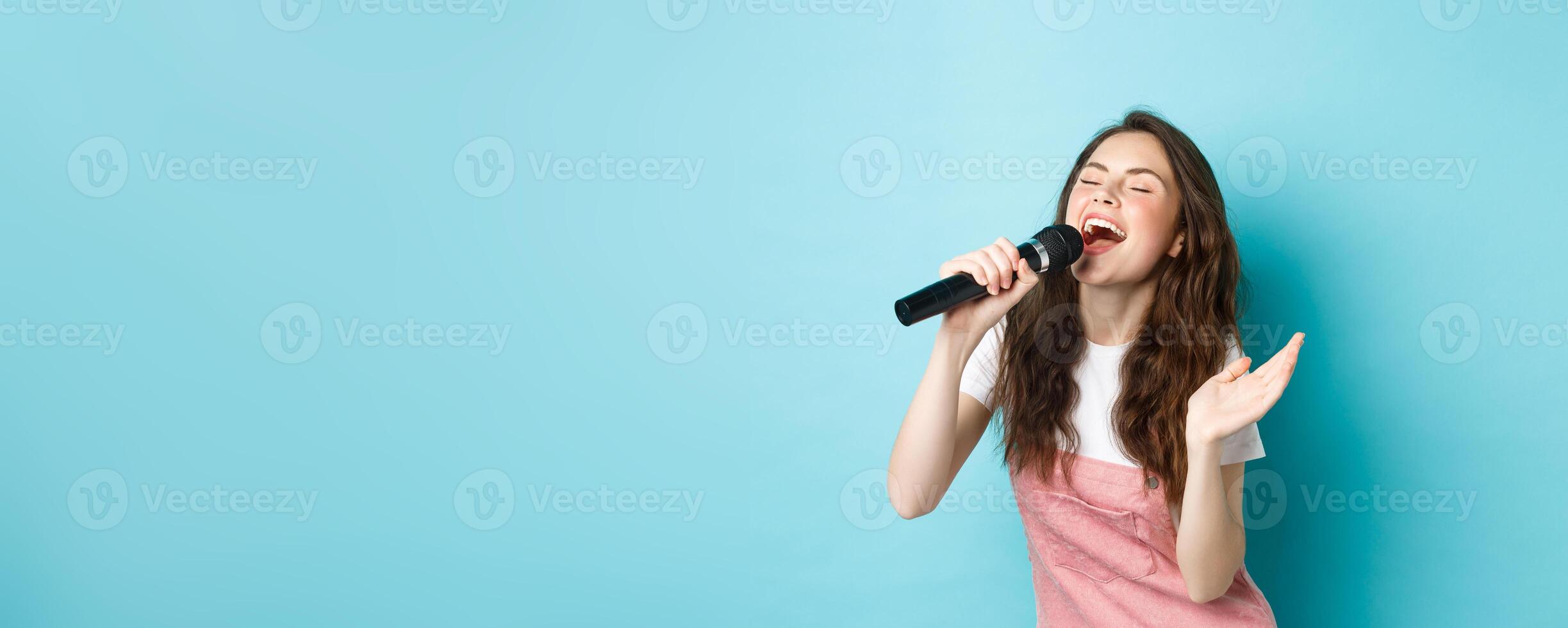 Carefree beautiful woman perform song, singing in microphone with passion, playing karaoke, standing over blue background photo