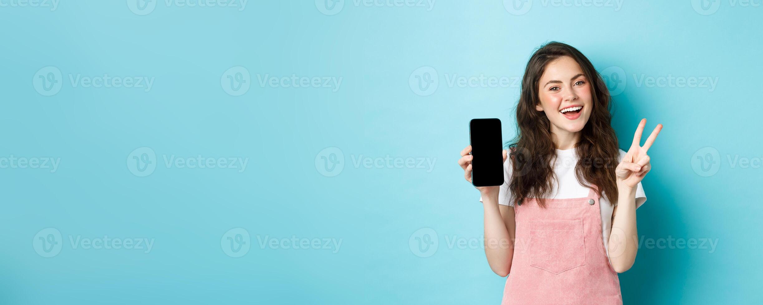 Cute young woman smiling and showing v-sign with empty smartphone screen, demonstrate app or mobile store, standing against blue background photo