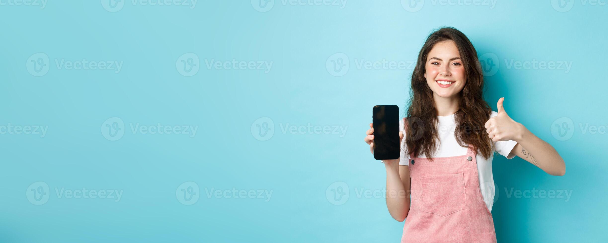 This is good. Smiling cheerful woman showng empty smartphone screen and thumb up to recommend app or online store, standing against blue background photo