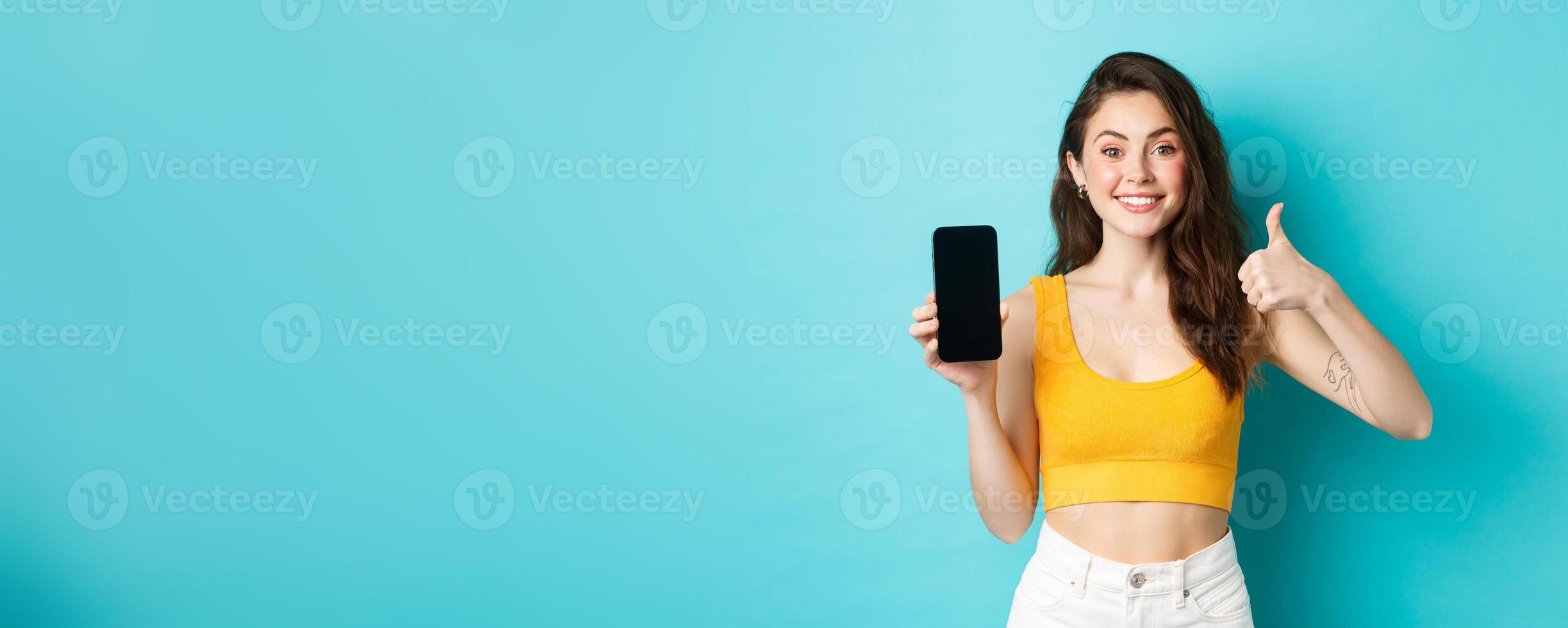 Portrait of stylish modern woman smiling pleased, recommending app, showing you logo banner on empty smartphone screen, making thumb up in approval, praise application, blue background photo