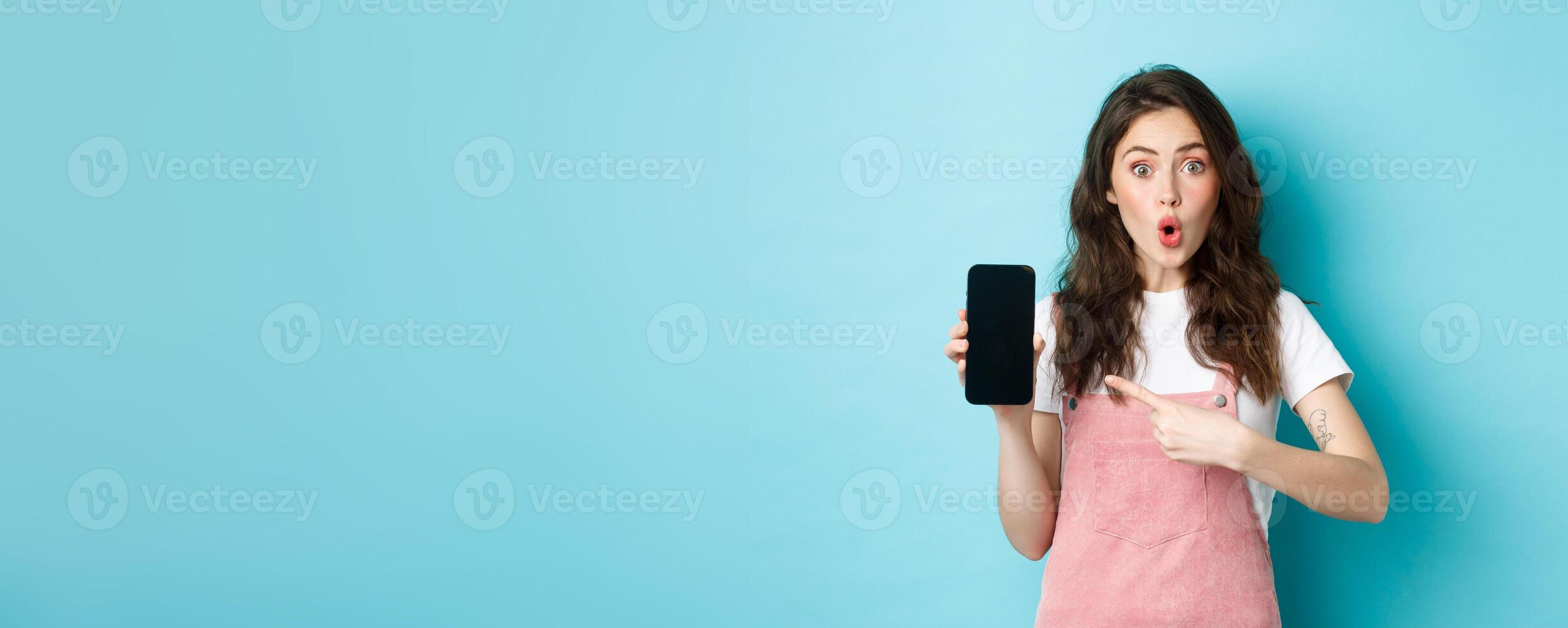 Impressed young modern woman pointing at smartphone screen, say wow and stare amazed at camera, showing awesome application, new online store, standing over blue background photo