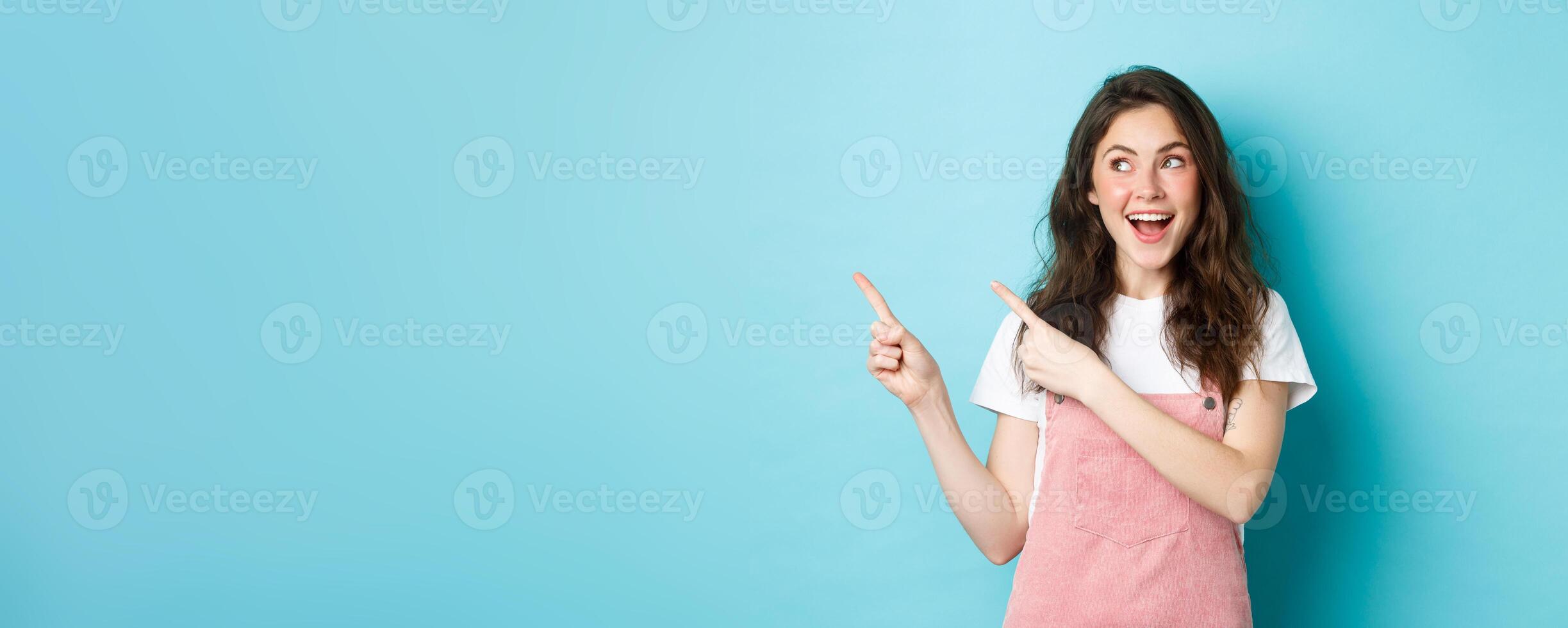 Portrait of happy smiling female model with glamour blush, pointing fingers left and looking at promo offer, showing an advertisement copy space, blue background photo