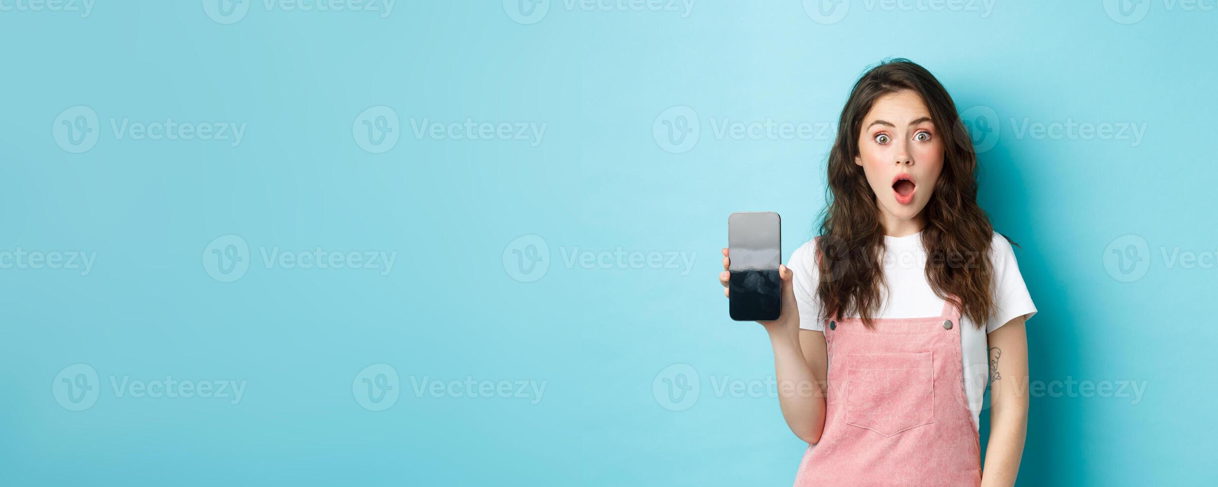 Shocked beautiful girl drop jaw and show empty smartphone screen, demonstrate your app or online store on blank phone display, standing against blue background photo