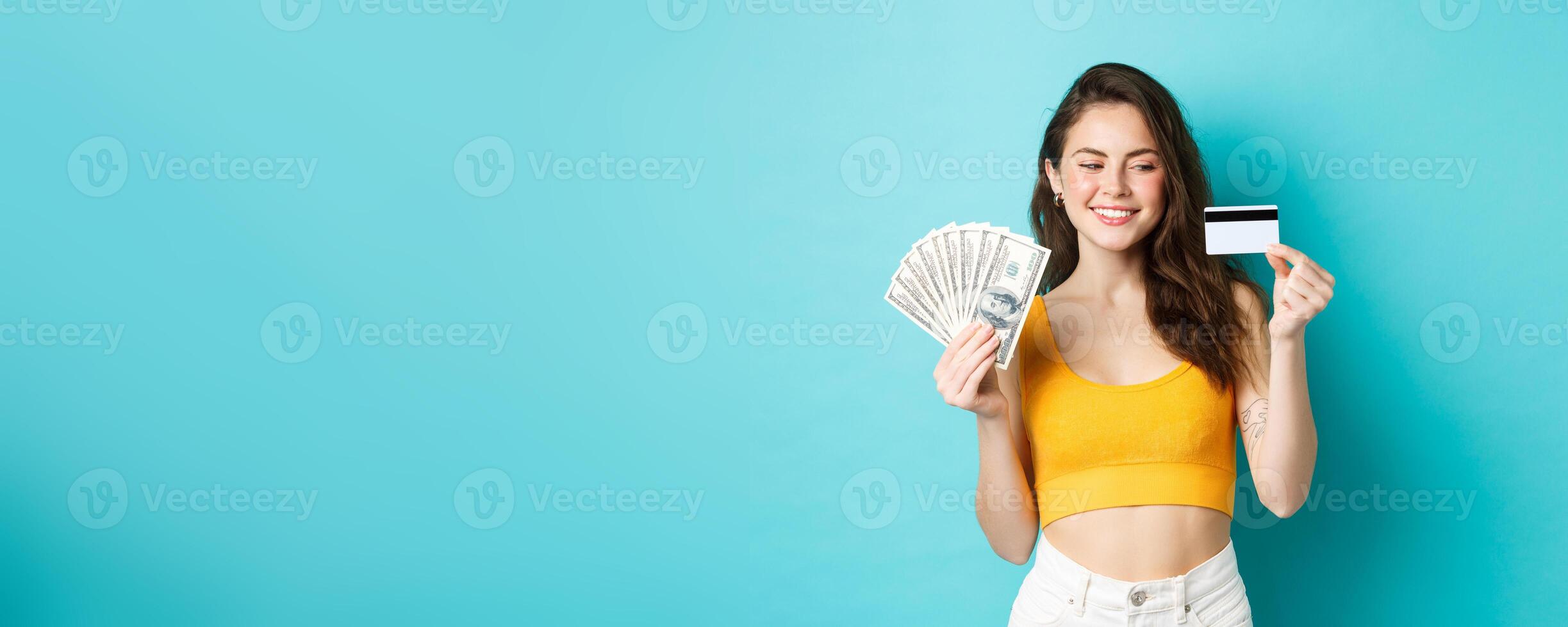 Good-looking smiling girl showing money but looking at plastic credit card with determined and pleased face, standing over blue background photo