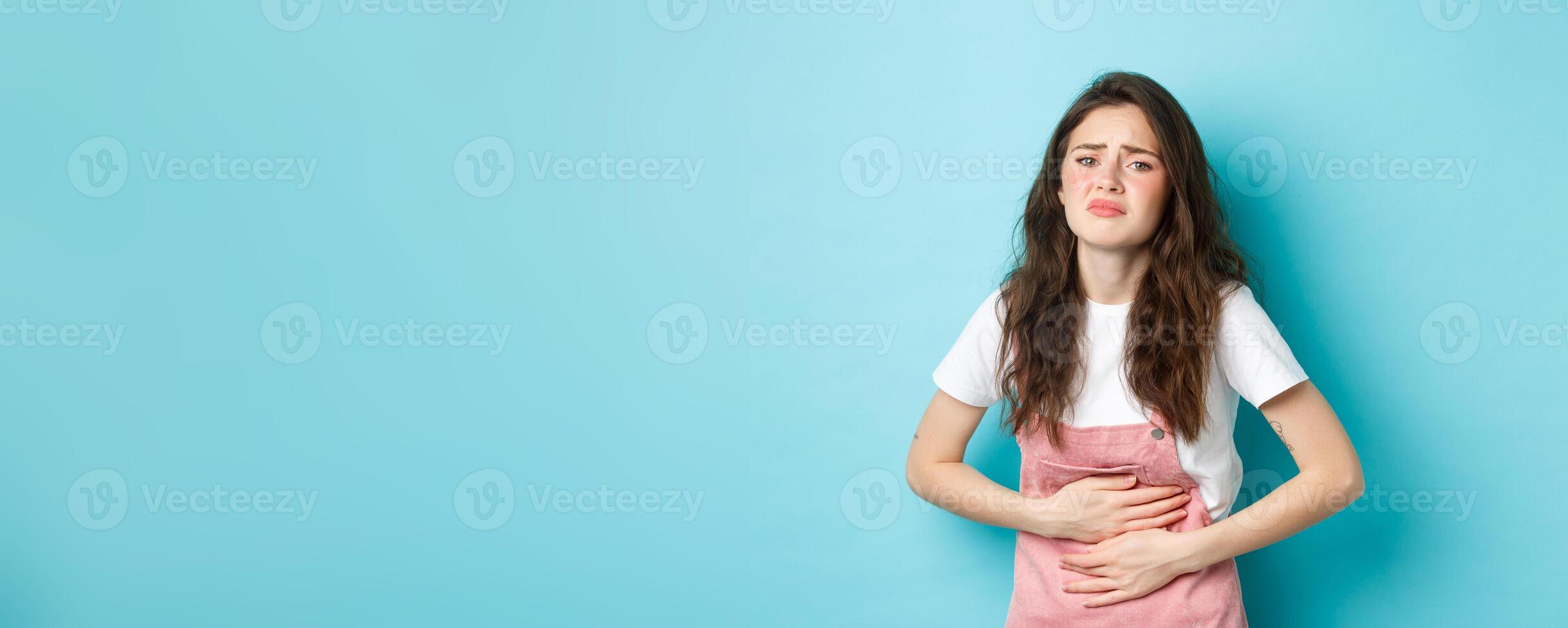 Image of sad young woman frowning, holding hands on stomach, having ache, painful menstrual cramps, standing against blue background photo