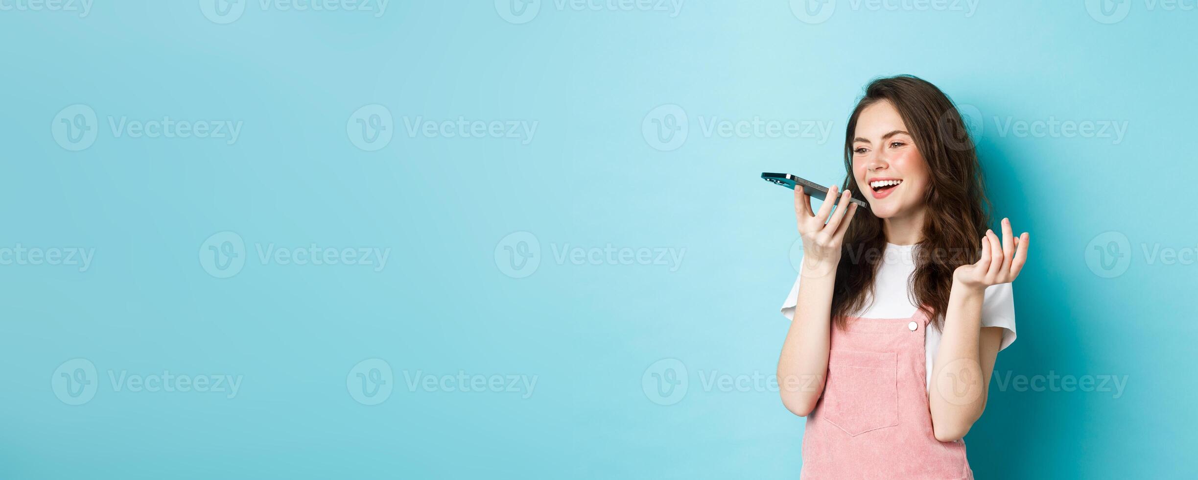 Portrait of smiling woman holding phone near lips and talking, using app translator on smartphone or recording voice message, speaking with speakerphone, standing over blue background photo