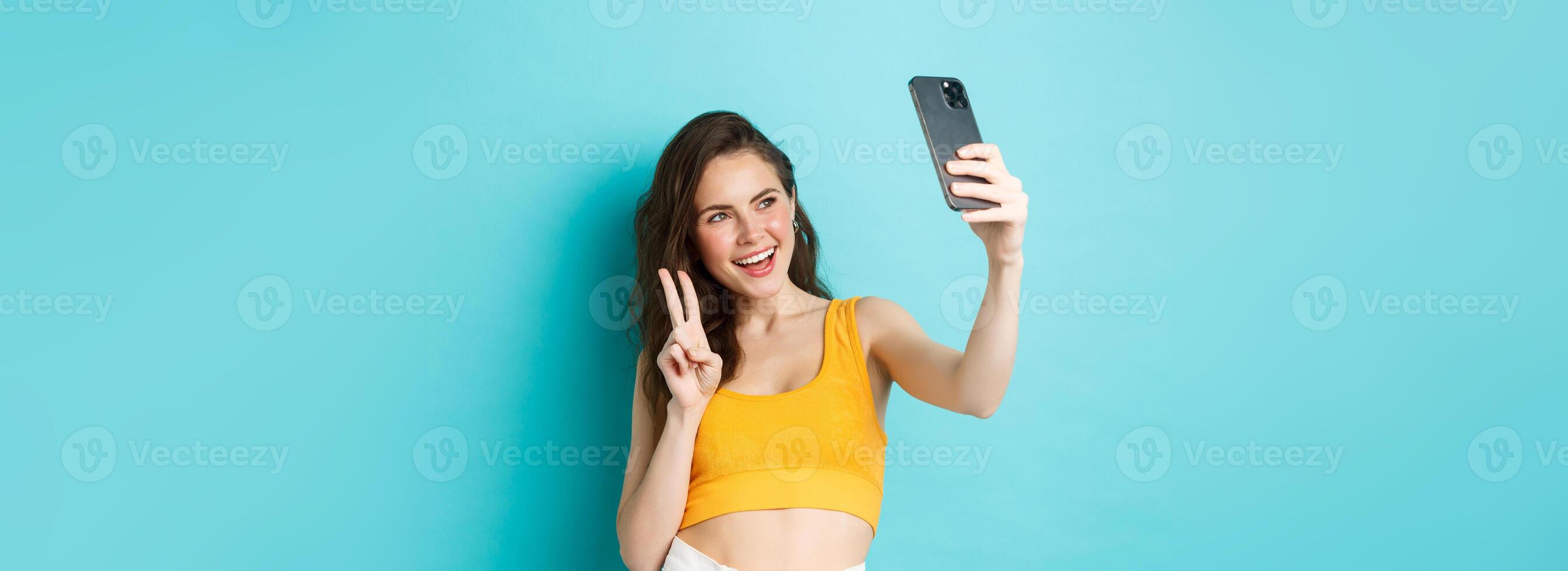 Technology and lifestyle concept. Stylish young woman taking selfie in her summer clothes, showing v-sign at smartphone camera, standing over blue background photo