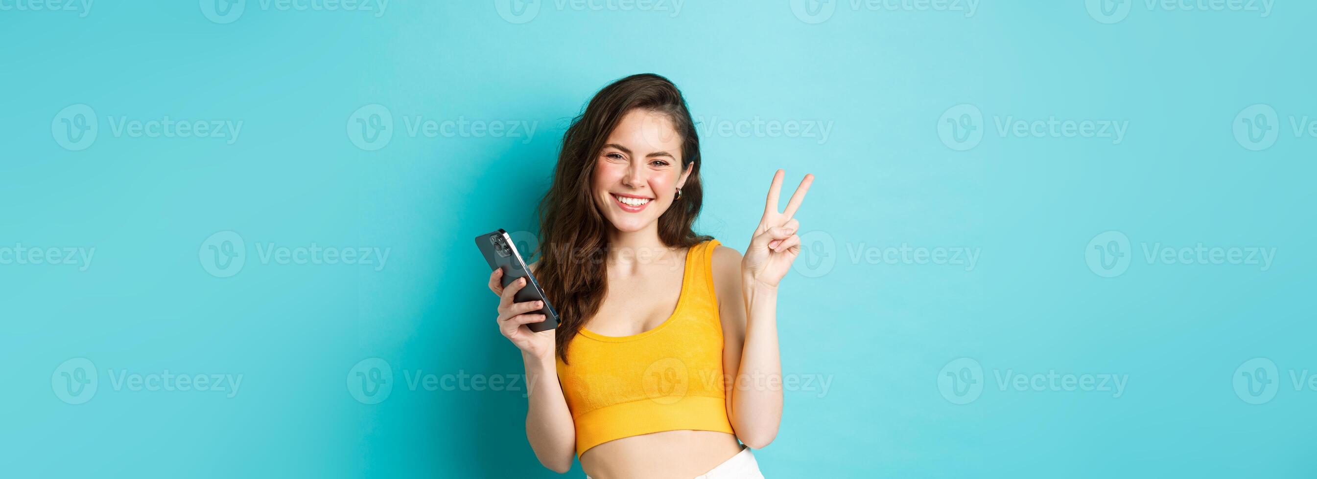 Technology and lifestyle concept. Beautiful cheerful girl sending positive vibes, smiling and showing peace sign, using cellphone, standing against blue background photo