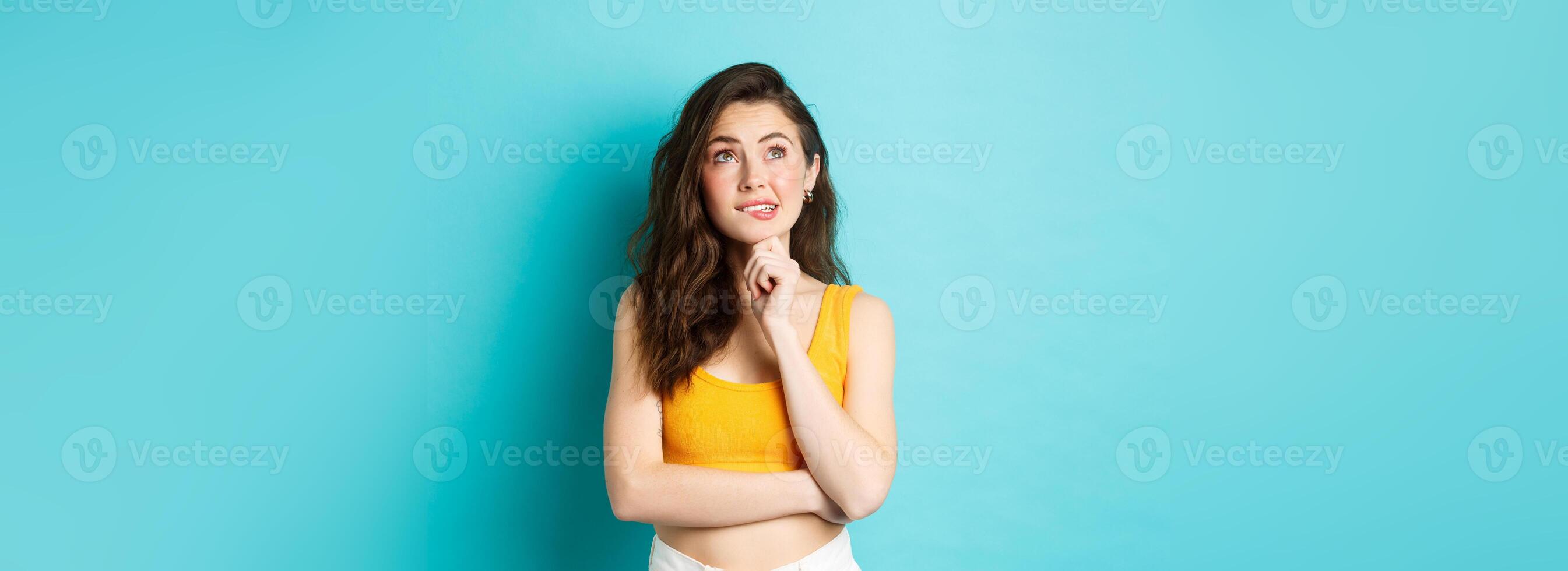 Pretty female model feeling indecisive, hesitating while making choice, looking at upper left corner and thinking, touching chin, standing over blue background photo