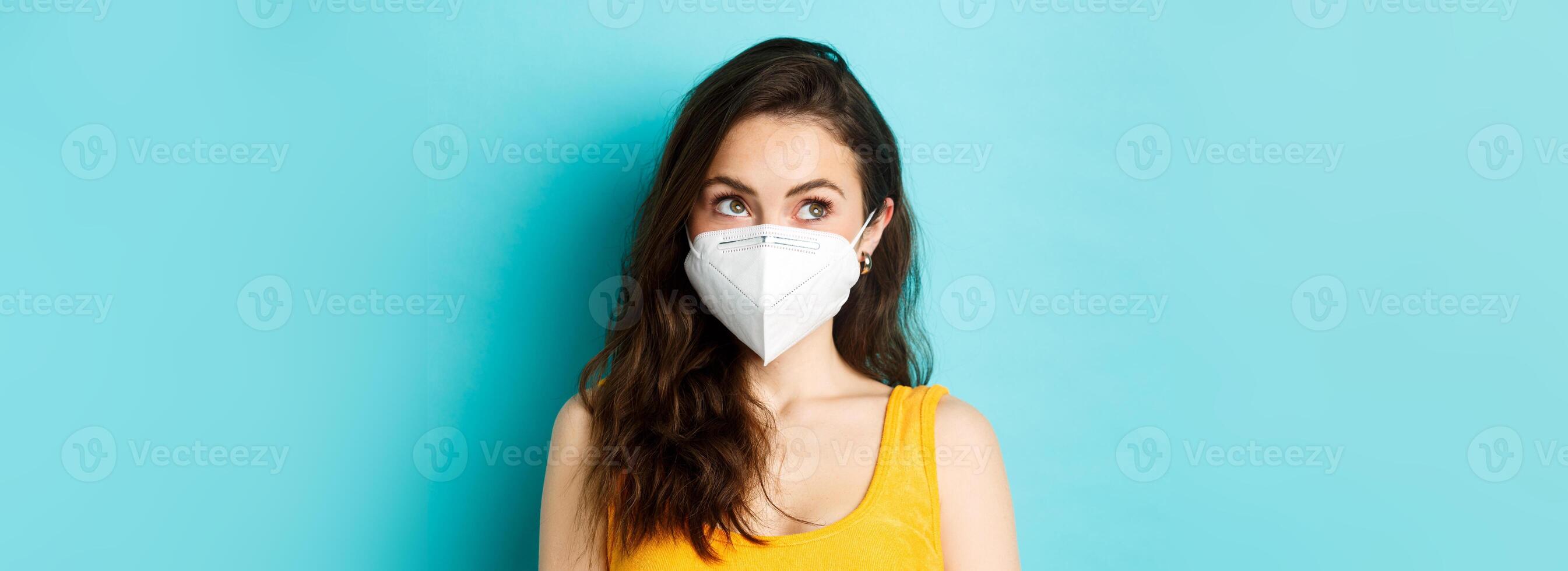 Covid-19, coronavirus and social distancing. Close up portrait of young modern woman in respirator, looking at upper left corner logo, standing against blue background photo