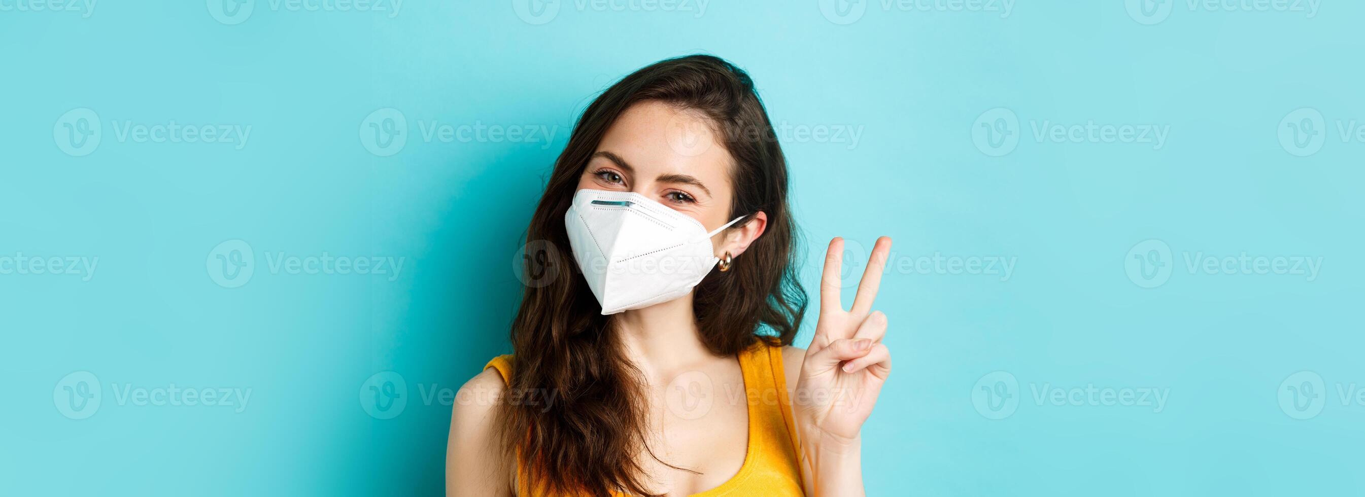 Covid-19, coronavirus and social distancing. Young positive woman in respirator showing v-sign, standing against blue background photo