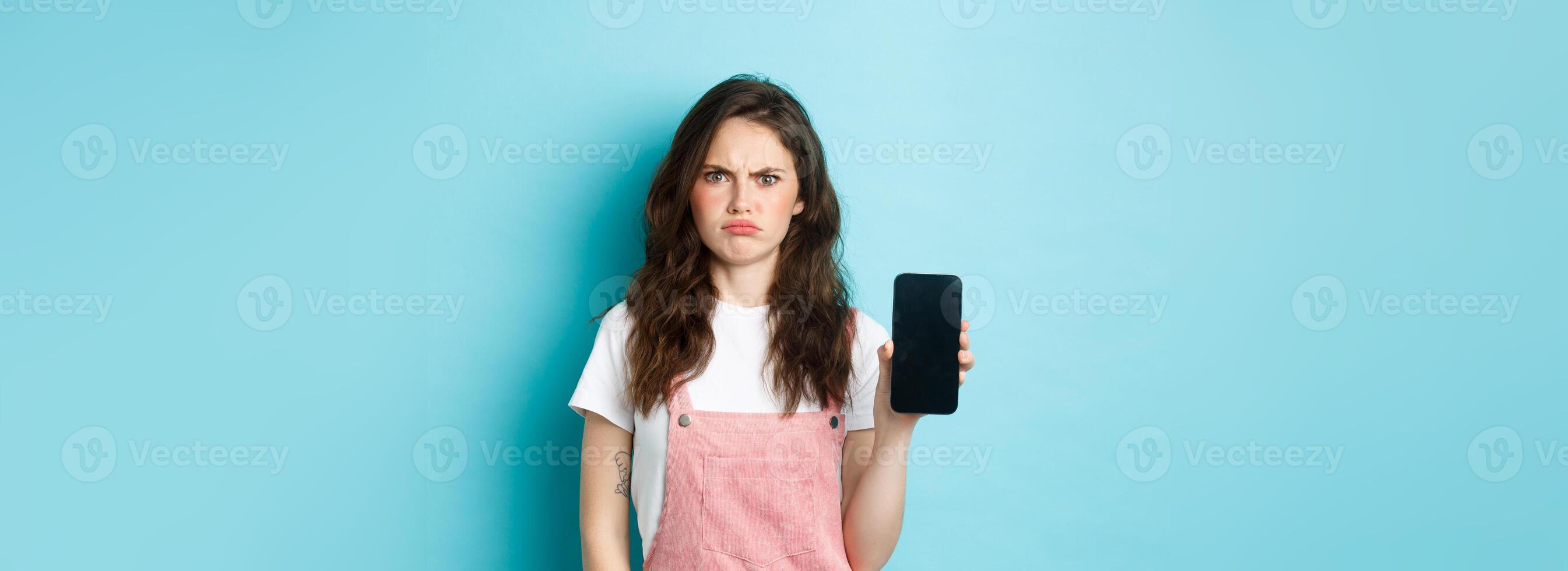 Disappointed girl frowning while showing empty smartphone screen, look displeased or angry, standing against blue background photo