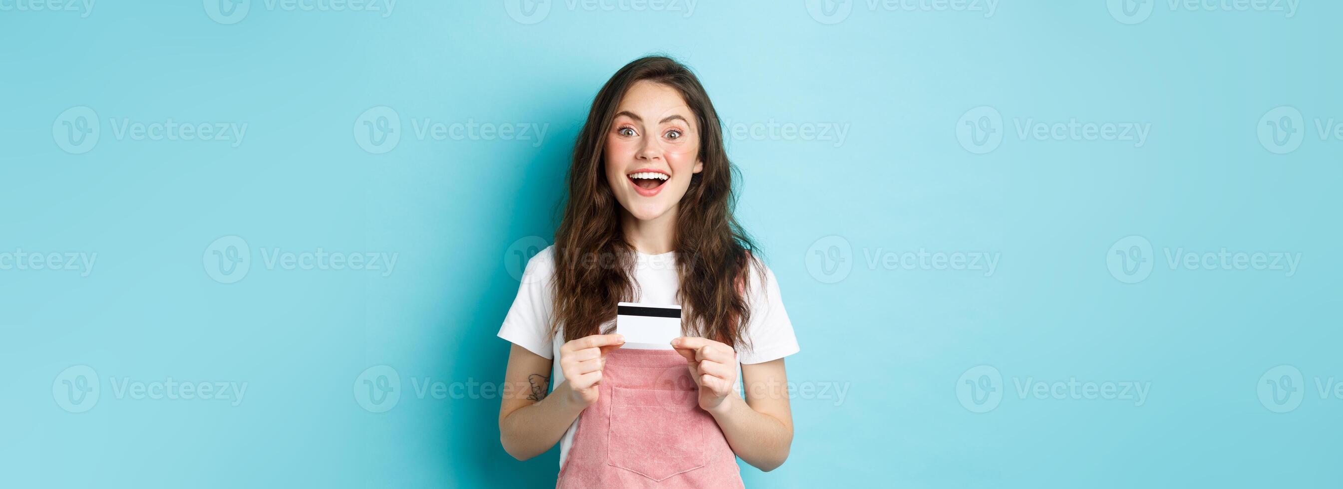 Excited beautiful female model showing plastic credit card, staring dreamy at camera, going shopping, standing over blue background photo