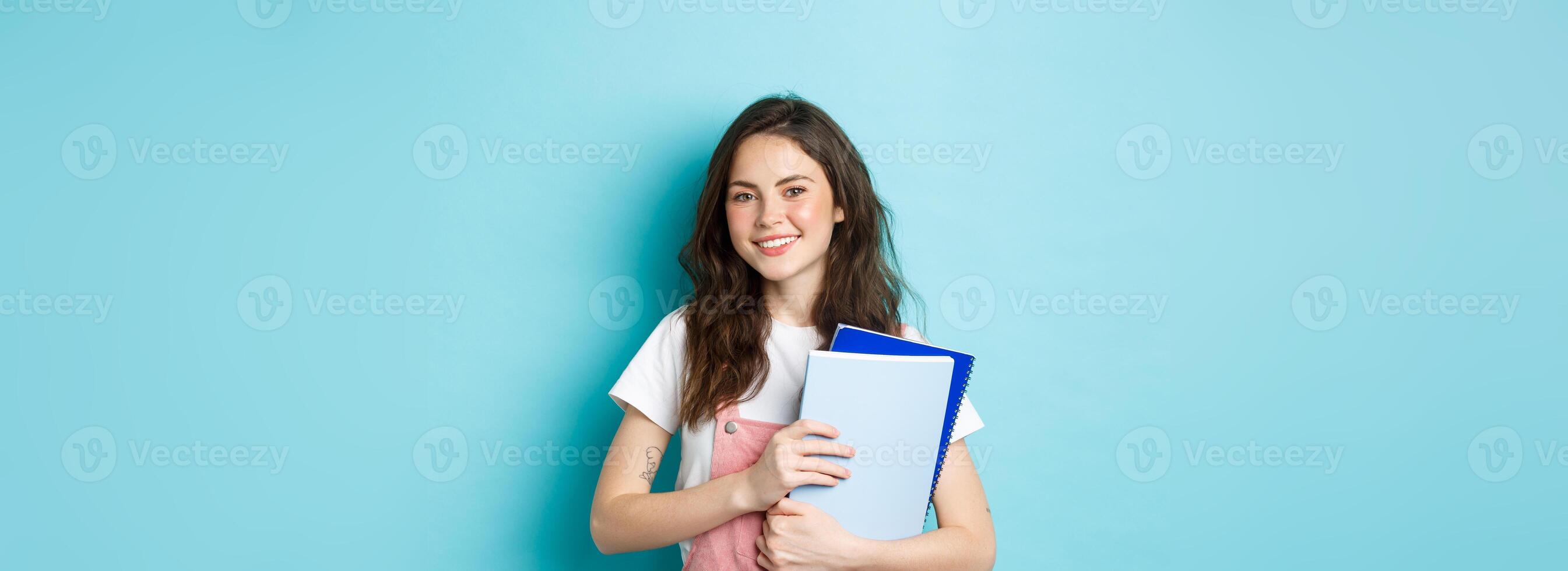 Cute smiling girl student holding notebooks and looking cheerful at camera, studying in college or university, standing over blue background photo