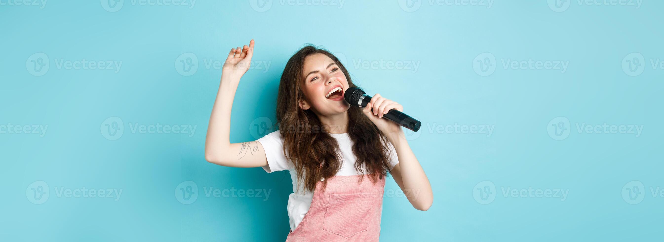 Happy young woman perform song, singer holding microphone, dancing and singing at karaoke, standing over blue background photo