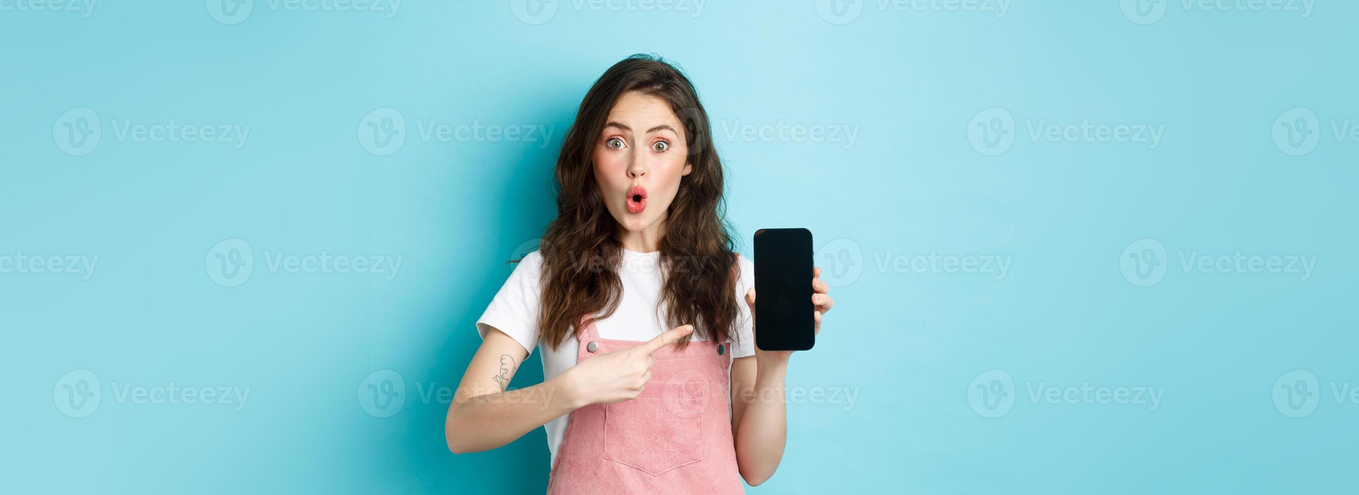 Impressed young modern woman pointing at smartphone screen, say wow and stare amazed at camera, showing awesome application, new online store, standing over blue background photo