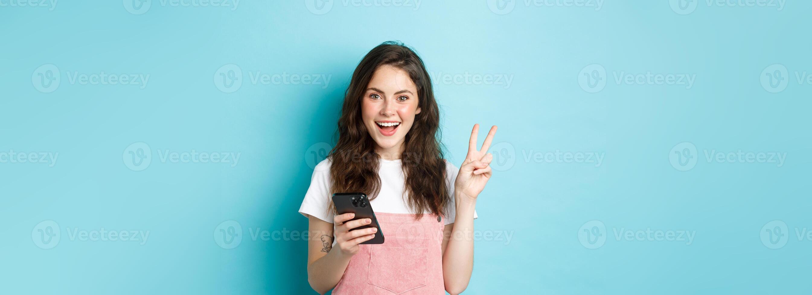 Online shopping. Positive smiling girl showing v-sign and looking happy at camera, using smartphone, holding mobile phone, standing against blue background photo