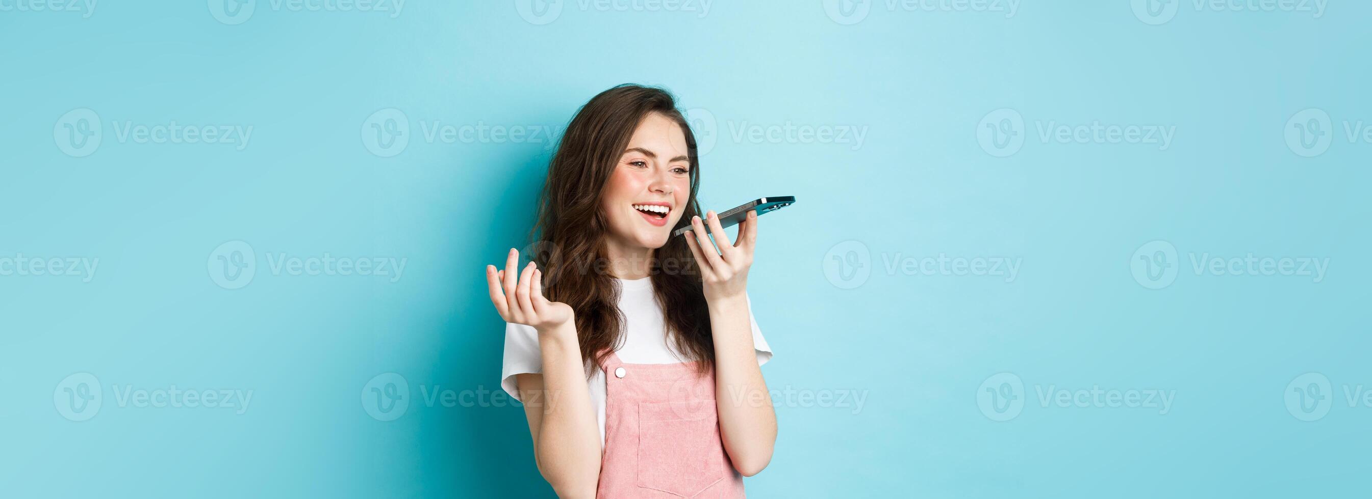 Portrait of smiling woman holding phone near lips and talking, using app translator on smartphone or recording voice message, speaking with speakerphone, standing over blue background photo