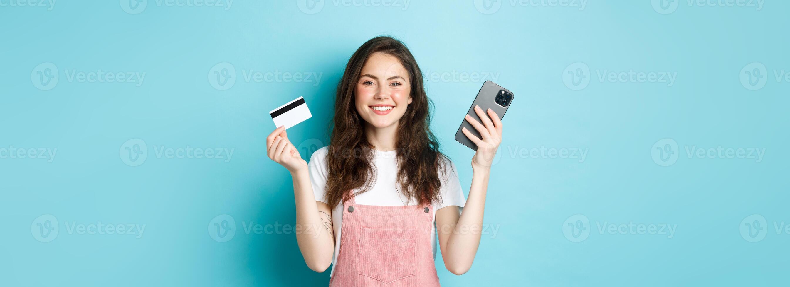 Smiling cute girl showing plastic credit card and smartphone, order something in store via mobile app, shop online, or pay for order, standing against blue background photo