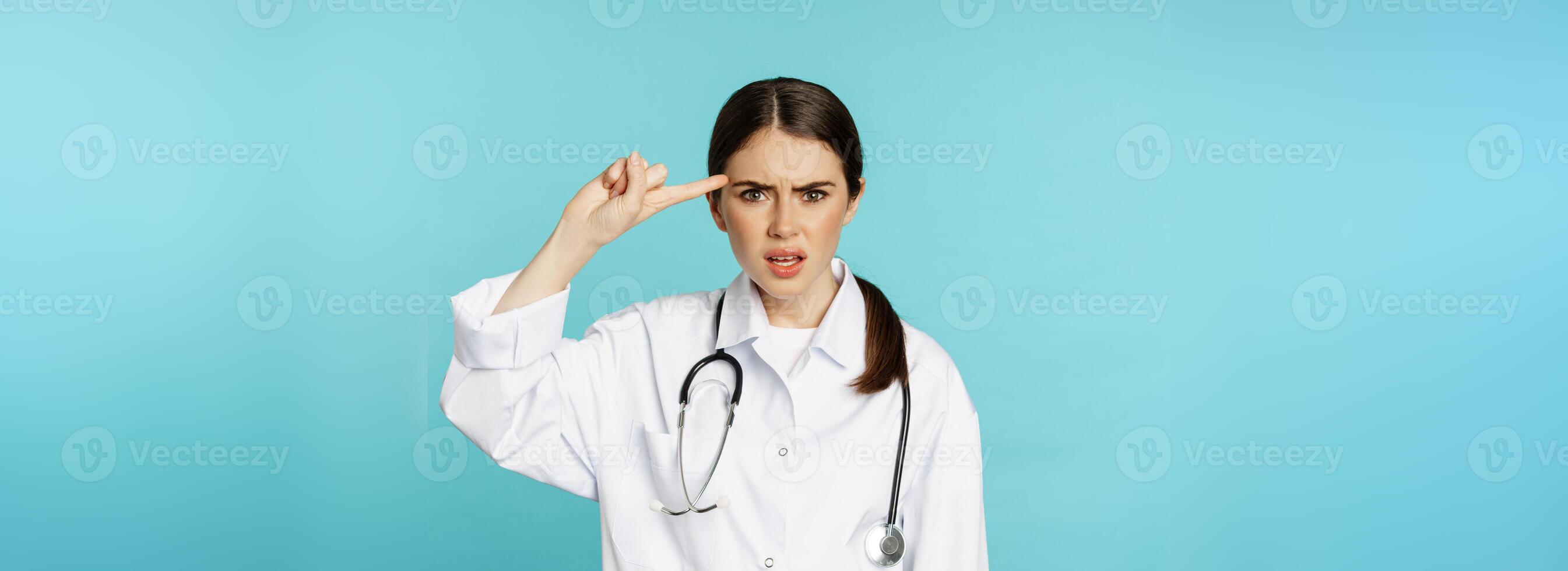 Annoyed woman doctor pointing finger at head, scolding someone stupid, crazy or strange, standing over torquoise background photo