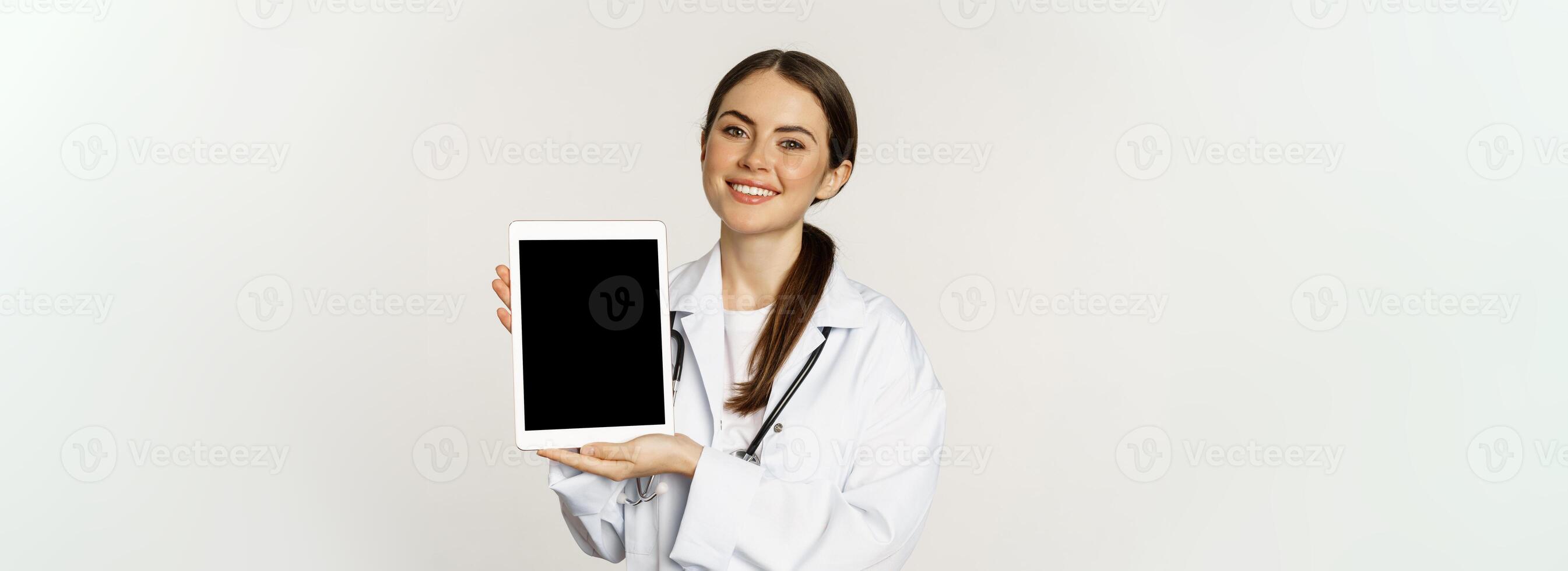 Online medical help, remote appointment. Smiling beautiful woman doctor showing digital tablet screen, demonstrating website logo, standing over white background photo