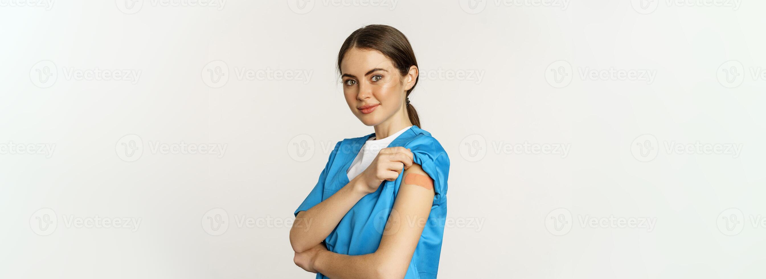 Female nurse, doctor medical worker got vaccinated, showing shoulder with patch after covid-19 vaccine, smiling pleased, standing over white background photo