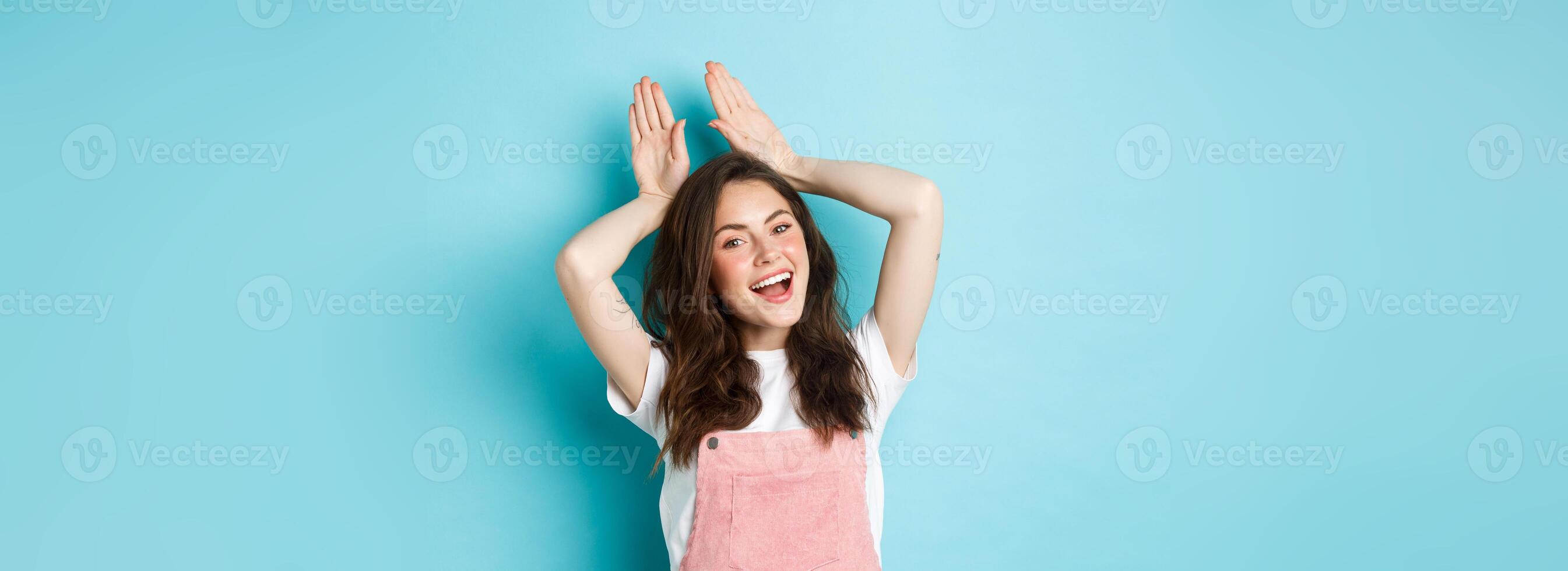 Beautiful girl mimic Easter bunny, showing rabbit ears with hands on head and smiling lovely, dancing happy, standing over blue background photo