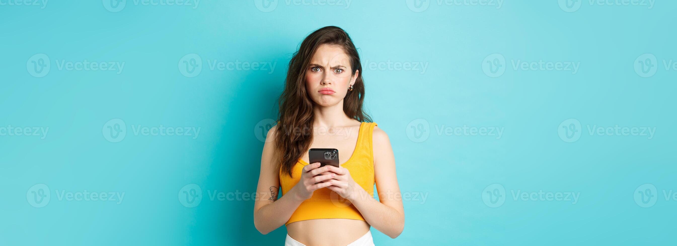 Confused and freak out young woman frowning, grimacing after reading strange message on smartphone, standing over blue background photo