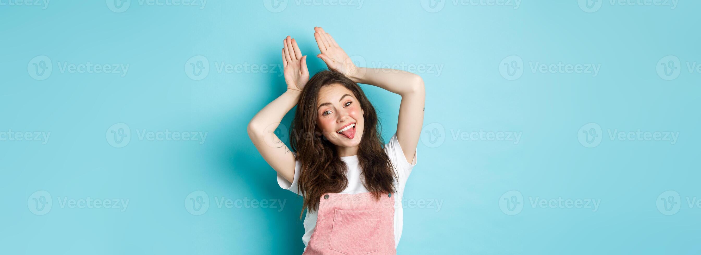 Portrait of beautiful glamour girl celebrating Easter, showing bunny rabbit ears with hands above head, smiling joyful, standing over blue background photo