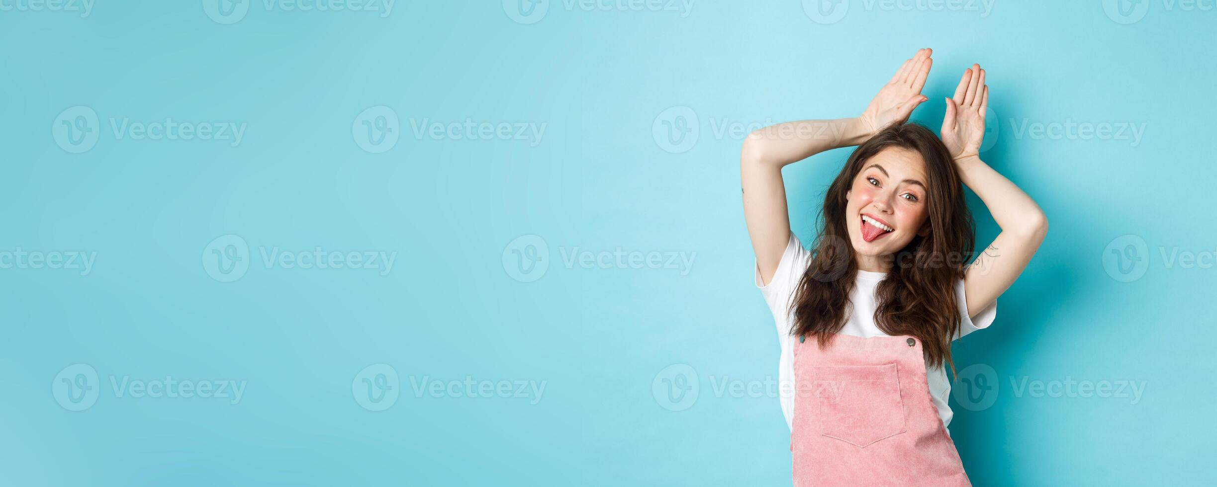 Portrait of beautiful glamour girl celebrating Easter, showing bunny rabbit ears with hands above head, smiling joyful, standing over blue background photo