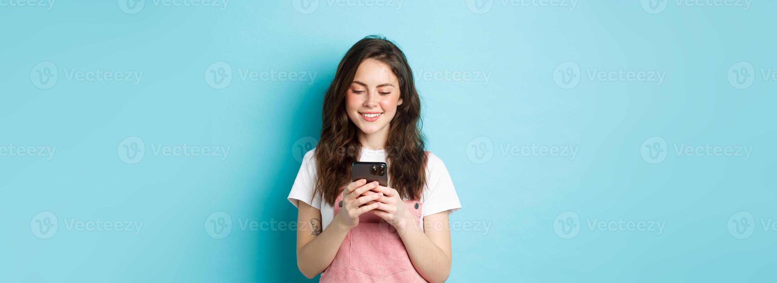 Portrait of young woman using smartphone, reading message or chatting in app, standing over blue background and smiling happy photo