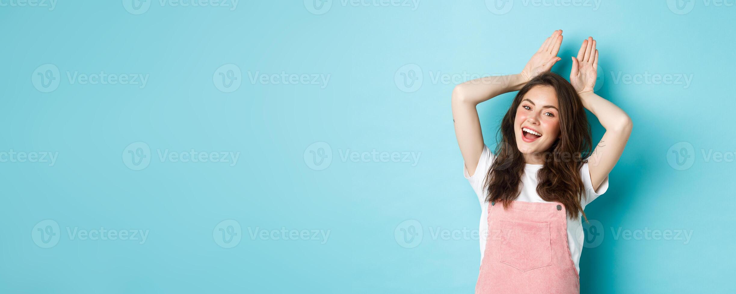 Beautiful girl mimic Easter bunny, showing rabbit ears with hands on head and smiling lovely, dancing happy, standing over blue background photo