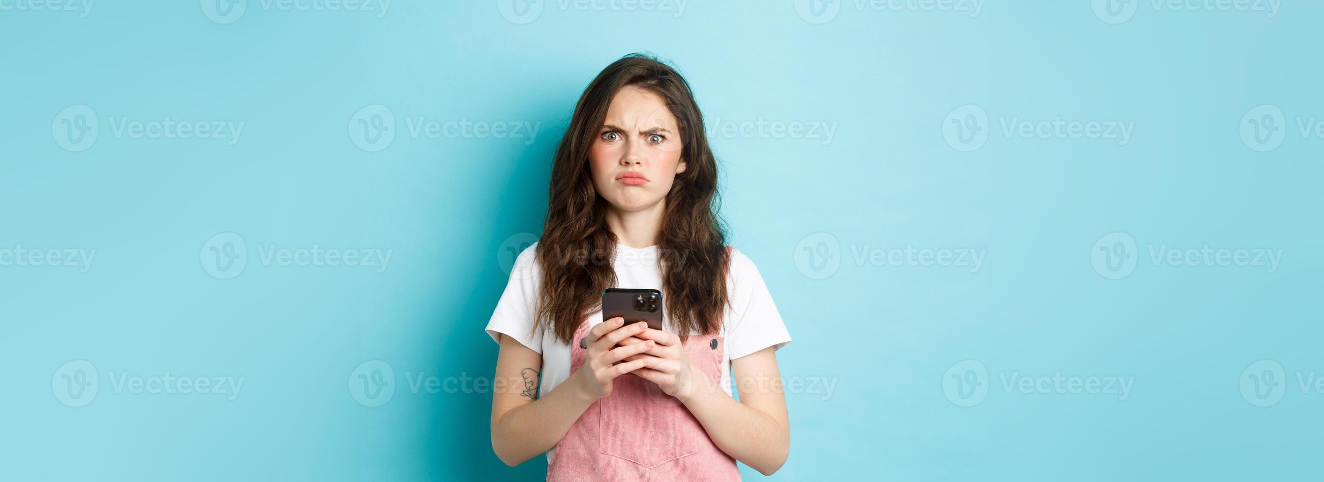 Disappointed girl frowning and grimacing displeased after using smartphone app, holding phone and express dislike, standing over blue background photo