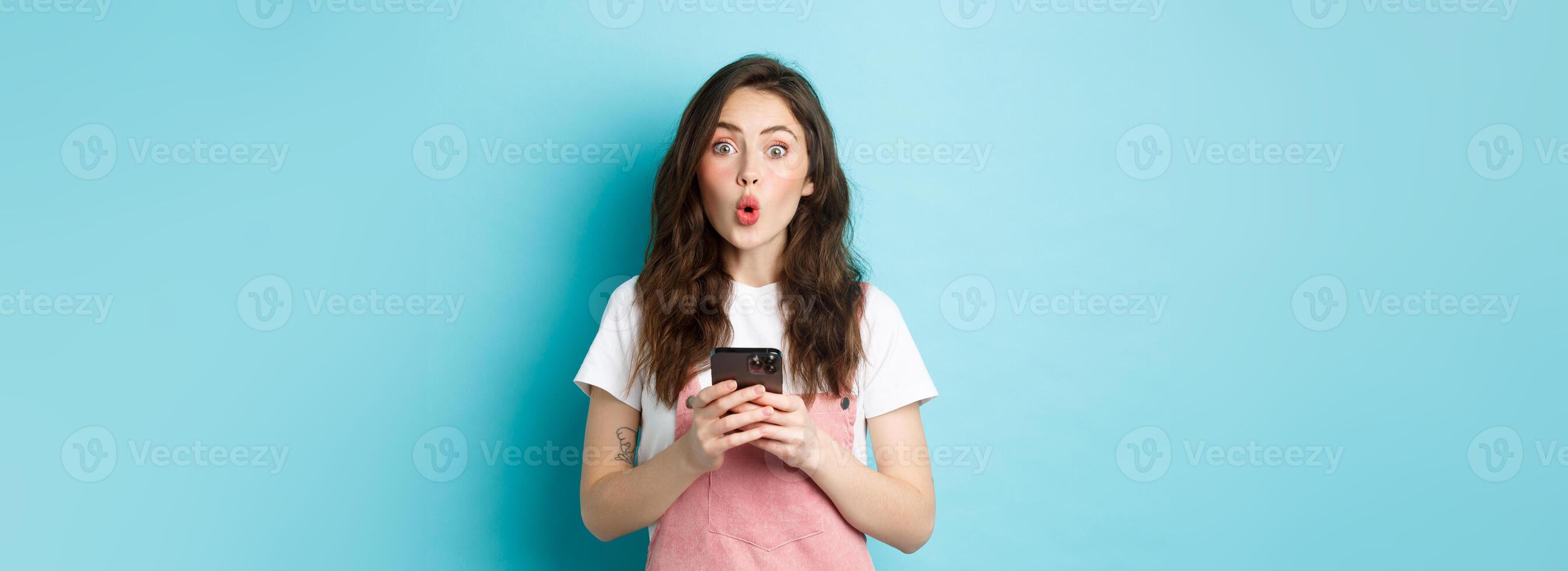 Portrait of brunette cute girl look surprised after using smartphone, holding mobile phone and say wow at camera with impressed face, blue background photo