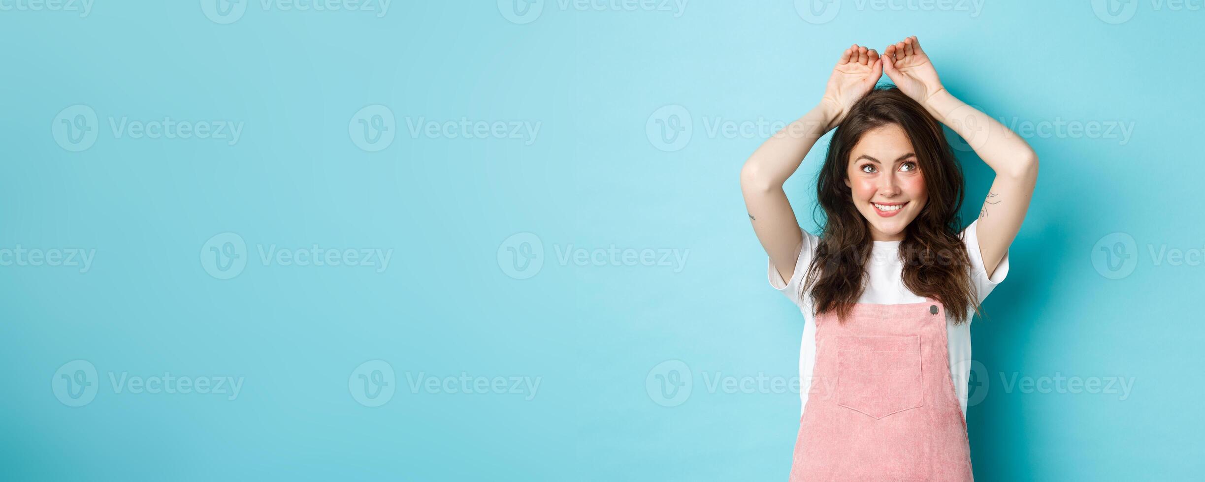 Cute young girl showing easter bunny gesture, making cute rabbig ears with hands on head and smiling silly, celebrating spring, standing over blue background photo