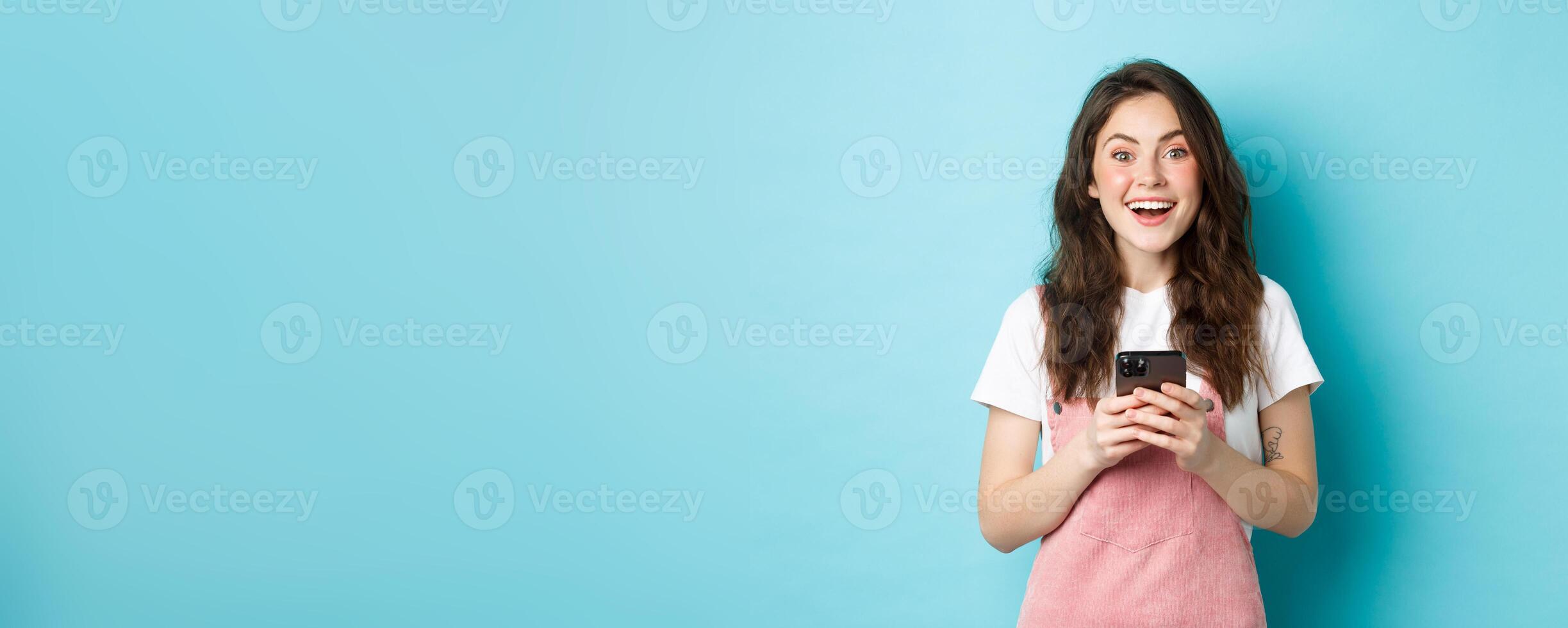 Portrait of excited brunette girl looking amazed at camera while using smartphone, holding mobile phone and smiling cheerful, standing against blue background photo