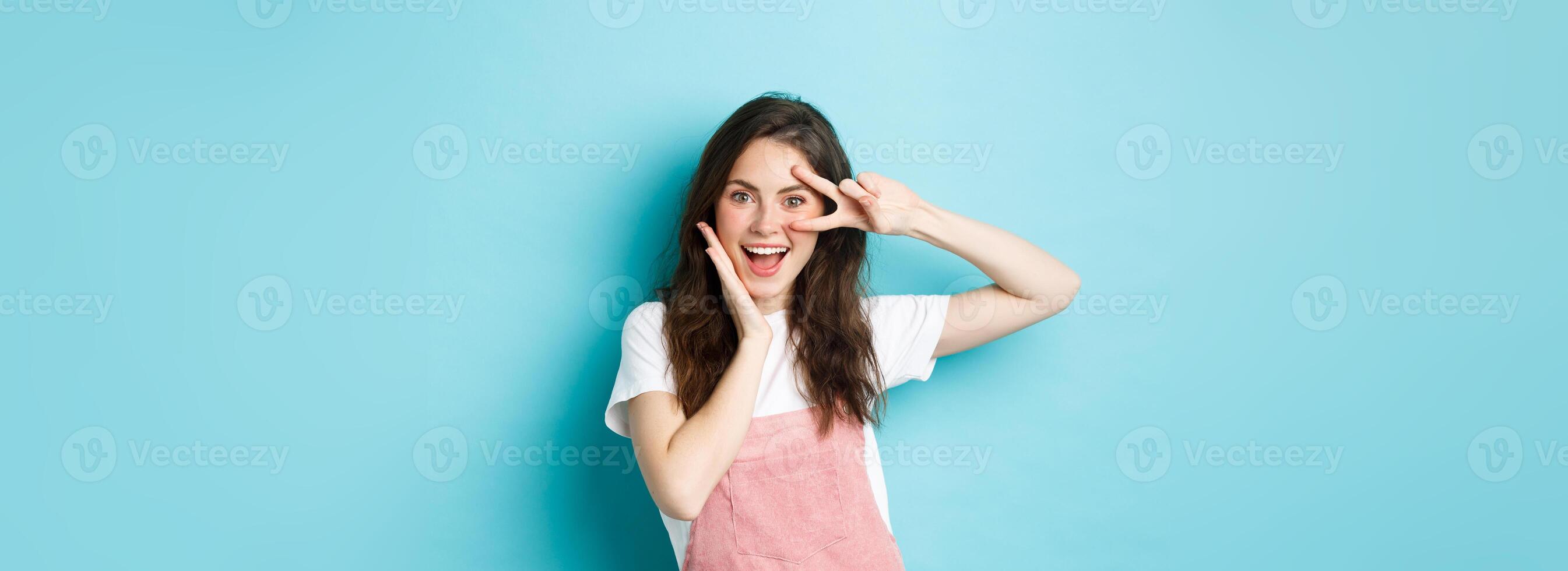 Portrait of excited attractive woman showing v-sign near eye, making kawaii gesture and smiling happy at camera, standing upbeat against blue background photo