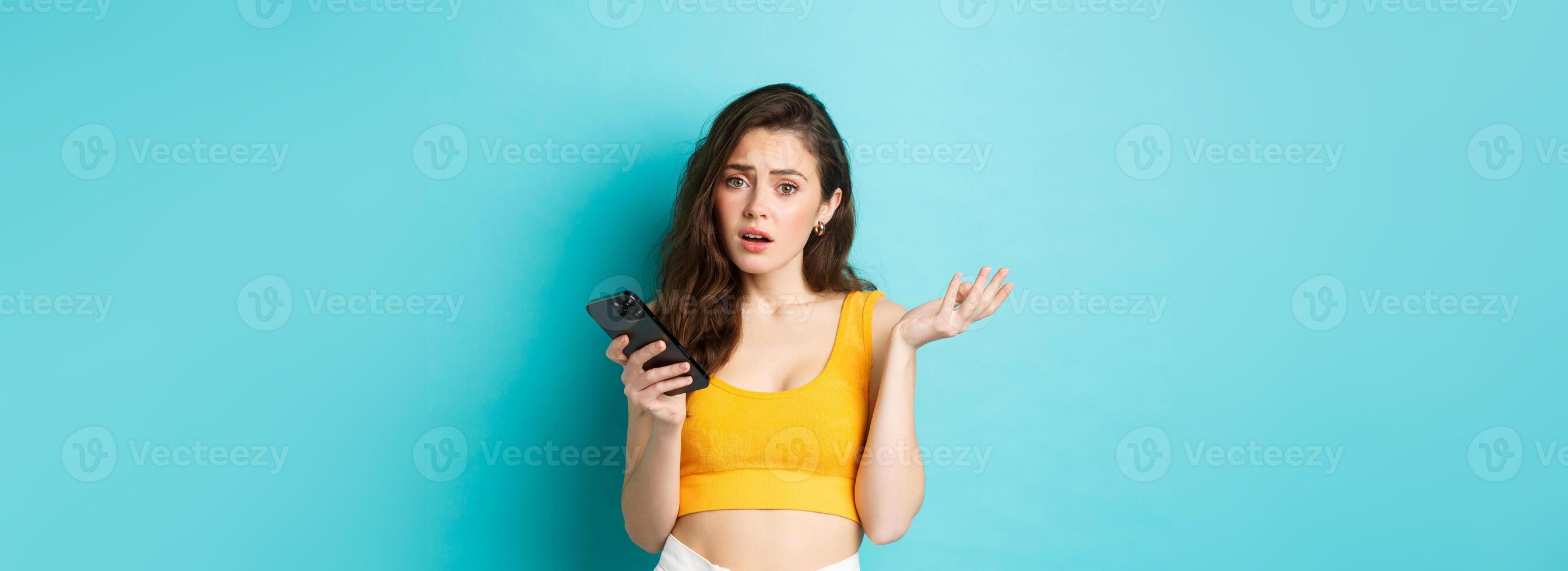 Technology and lifestyle concept. Glamour girl looks confused after reading smartphone screen, staring puzzled at camera, standing against blue background photo