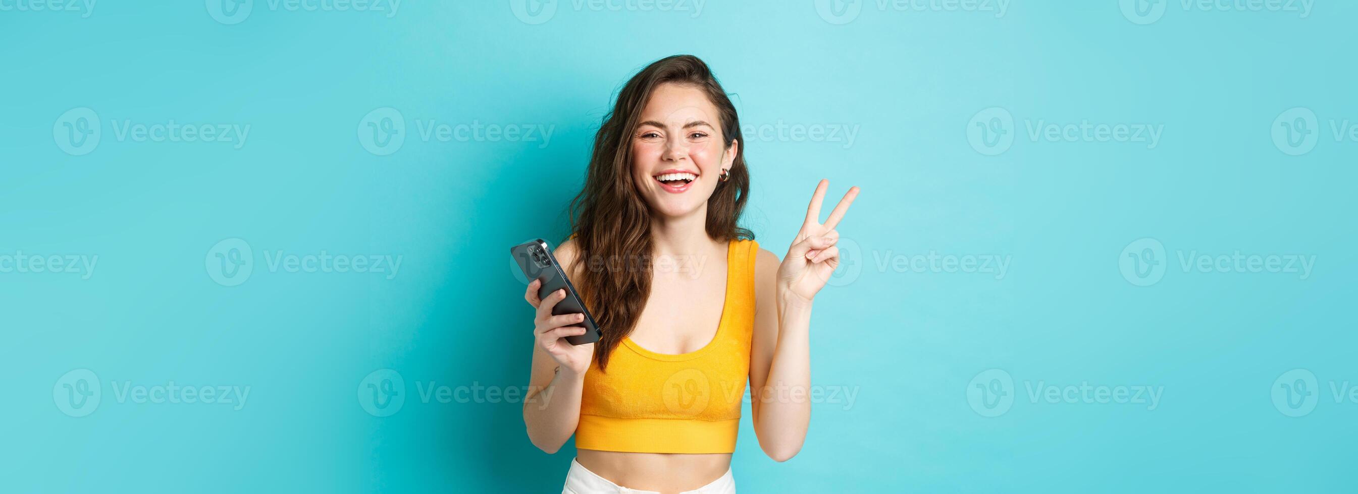 Technology and lifestyle concept. Happy attractive woman laughing, showing v-sign and using smartphone, chatting on mobile phone, standing over blue background photo