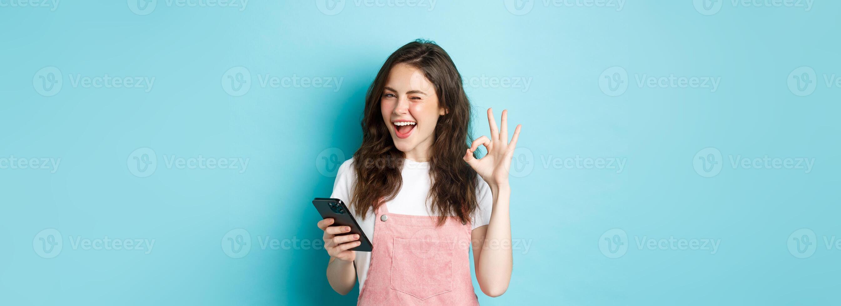 Online shopping. Cheerful cute girl winking at you, smiling and showing okay sign after using smartphone app, recommending internet shop or social media page, blue background photo