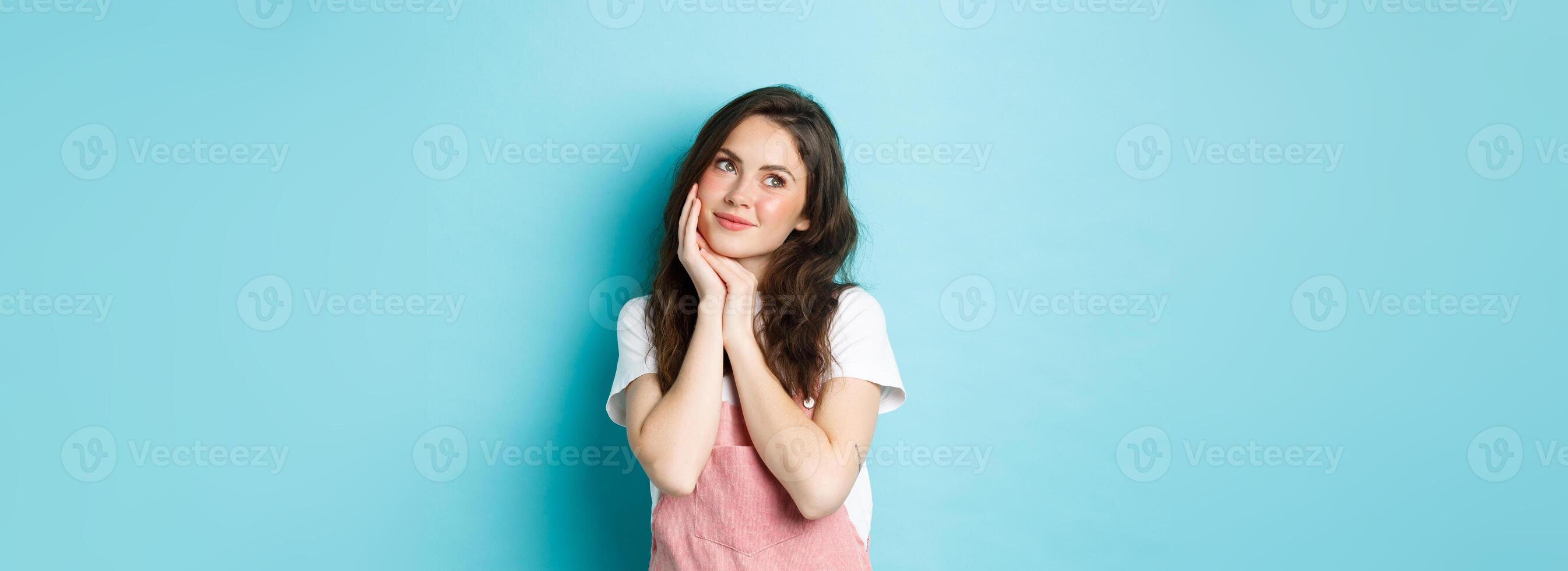 Portrait of beautiful girl dreaming, looking aside at logo banner and thinking, standing romantic against blue background, smiling and imaging something cute, blue background photo