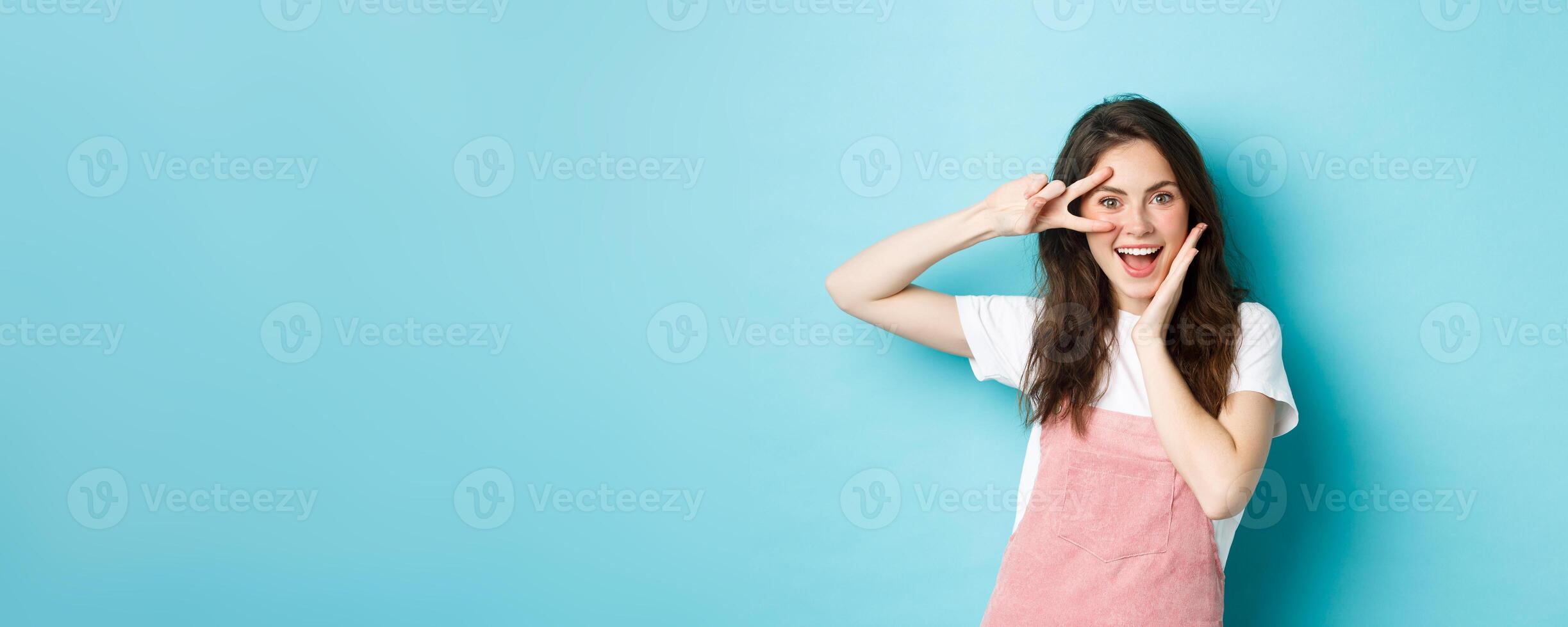 Portrait of excited attractive woman showing v-sign near eye, making kawaii gesture and smiling happy at camera, standing upbeat against blue background photo