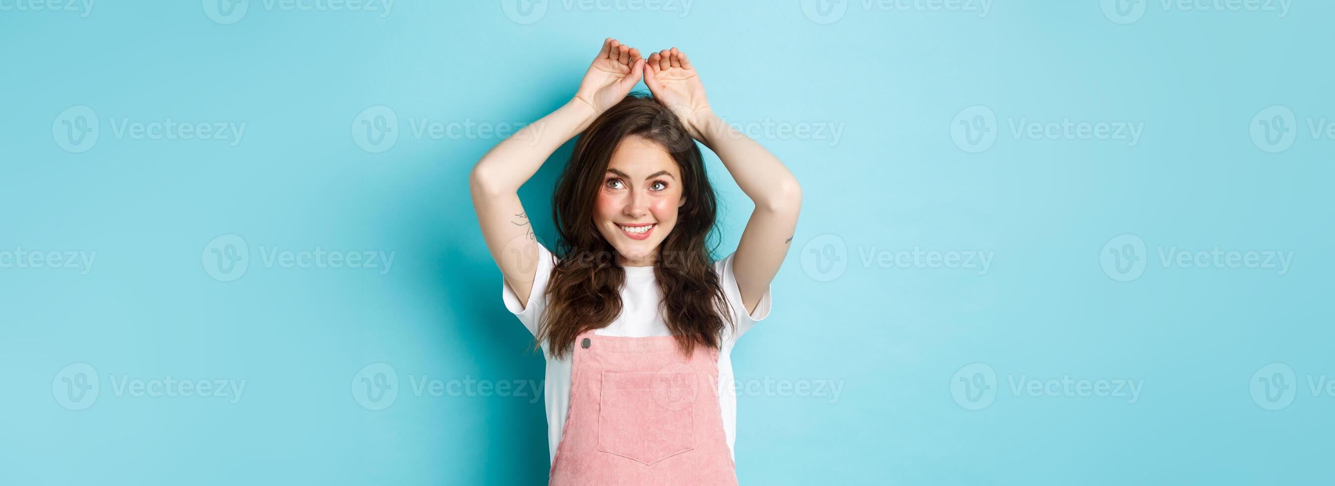 Cute young girl showing easter bunny gesture, making cute rabbig ears with hands on head and smiling silly, celebrating spring, standing over blue background photo