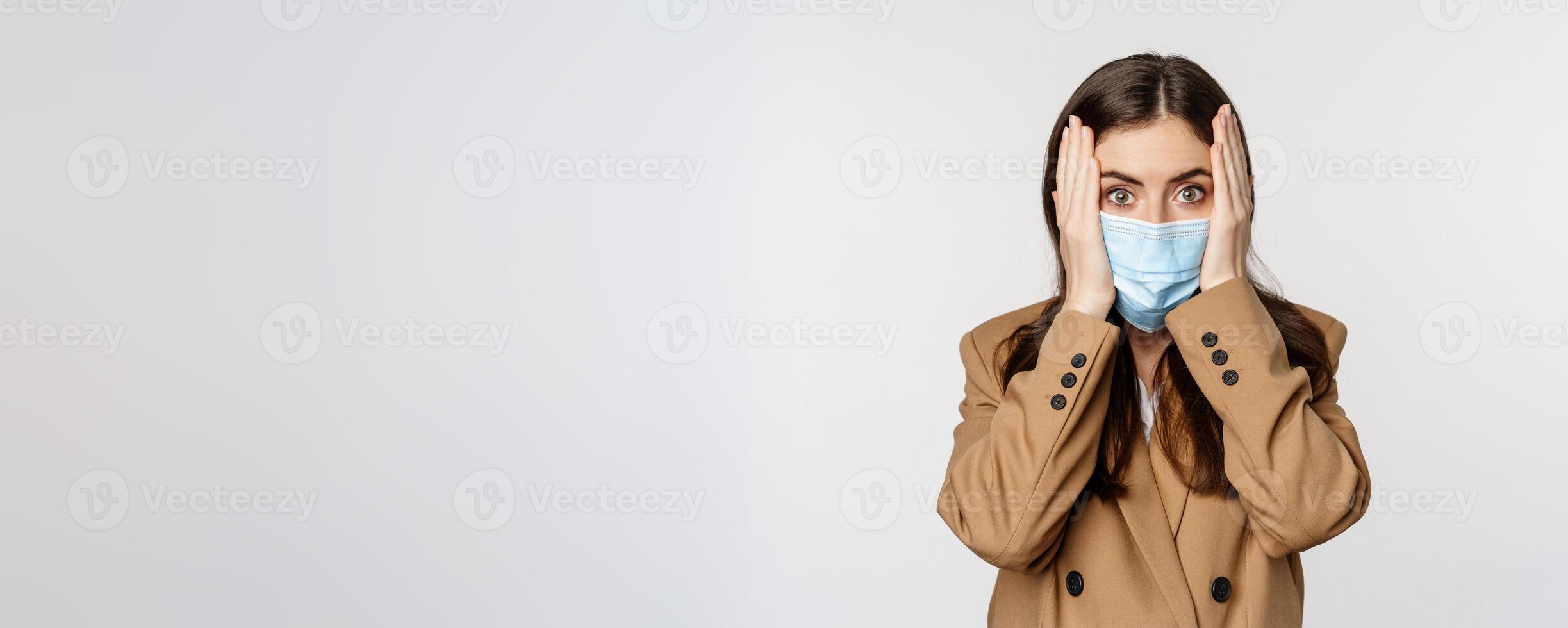 Covid-19 and people concept. Concerned, troubled office lady in medical face mask, holding hands on head and staring shocked at camera, white background photo