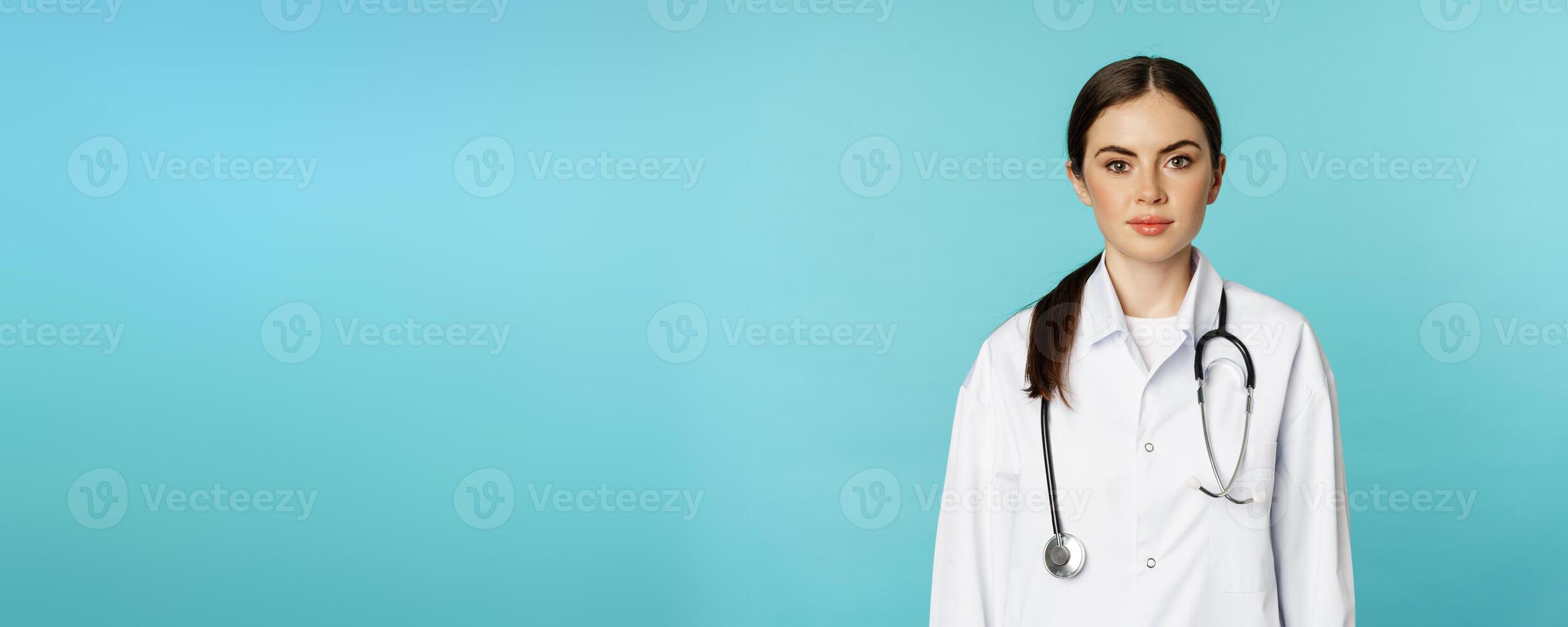 Medical staff and doctors concept. Young smiling female doctor, healthcare worker in white coat and stethoscope, looking confident, waiting patients, blue background photo