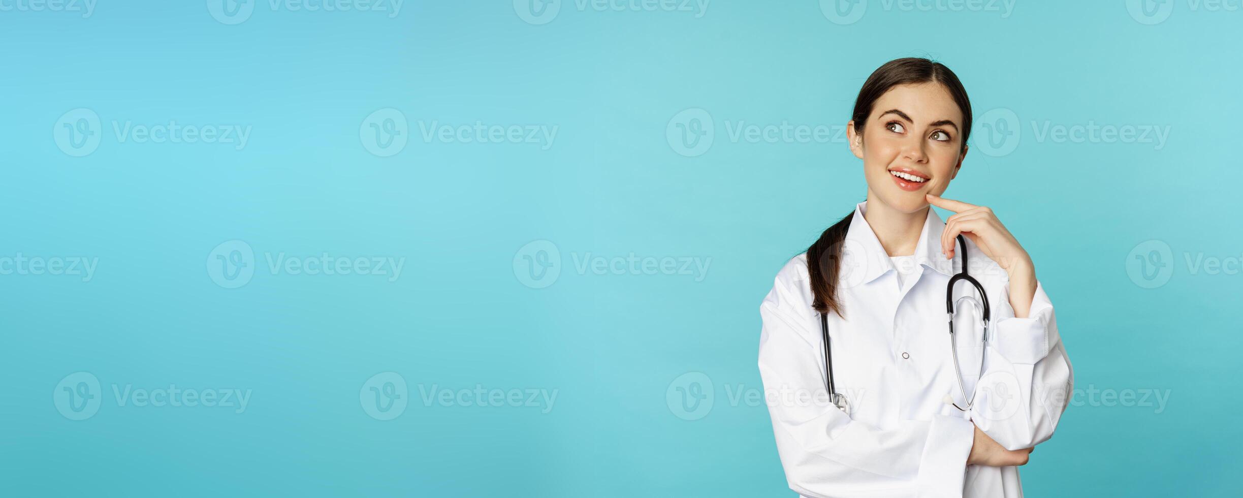 Young female doctor, hospital worker in white coat, thinking and looking away thoughtful, searching solution, standing over toquoise background photo