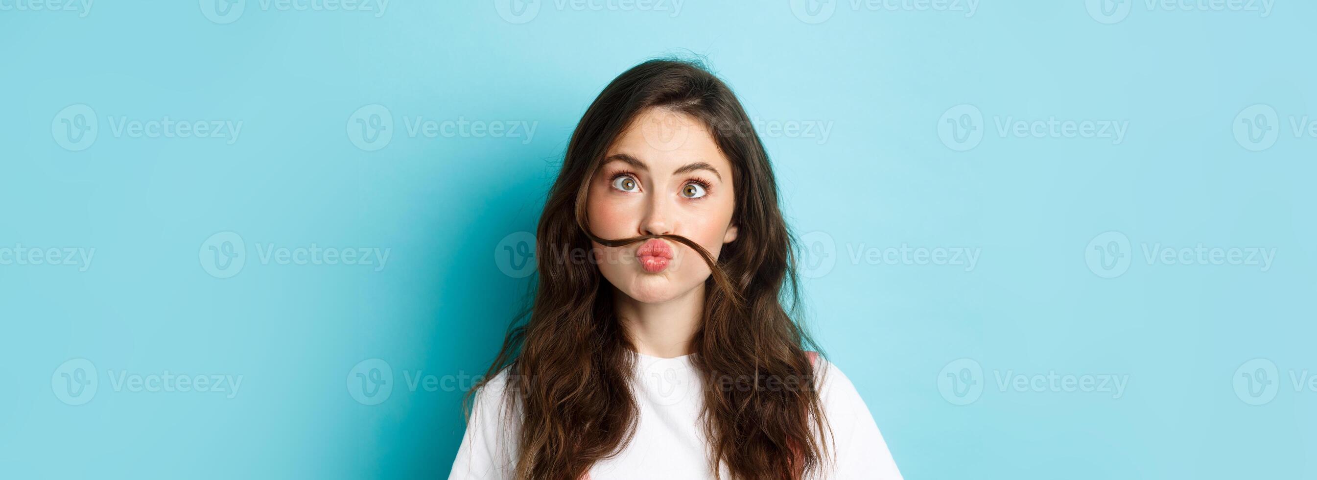 Close up portrait of funny silly girl playing, making squinting face and moustache with hair strand above lips, standing over blue background photo