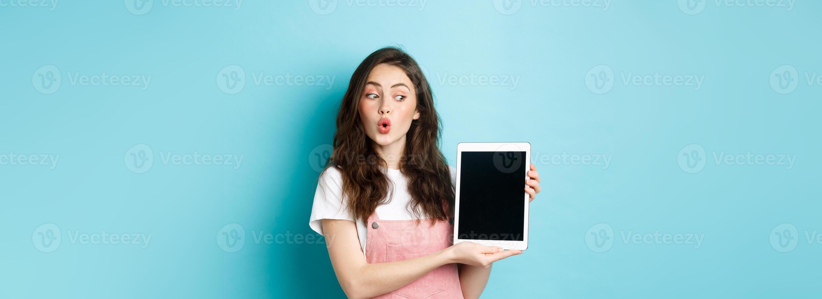 Intrigued glamour girl looking at digital tablet screen while showing it to you, standing amused with online promo offer, demonstrate something on display, blue background photo