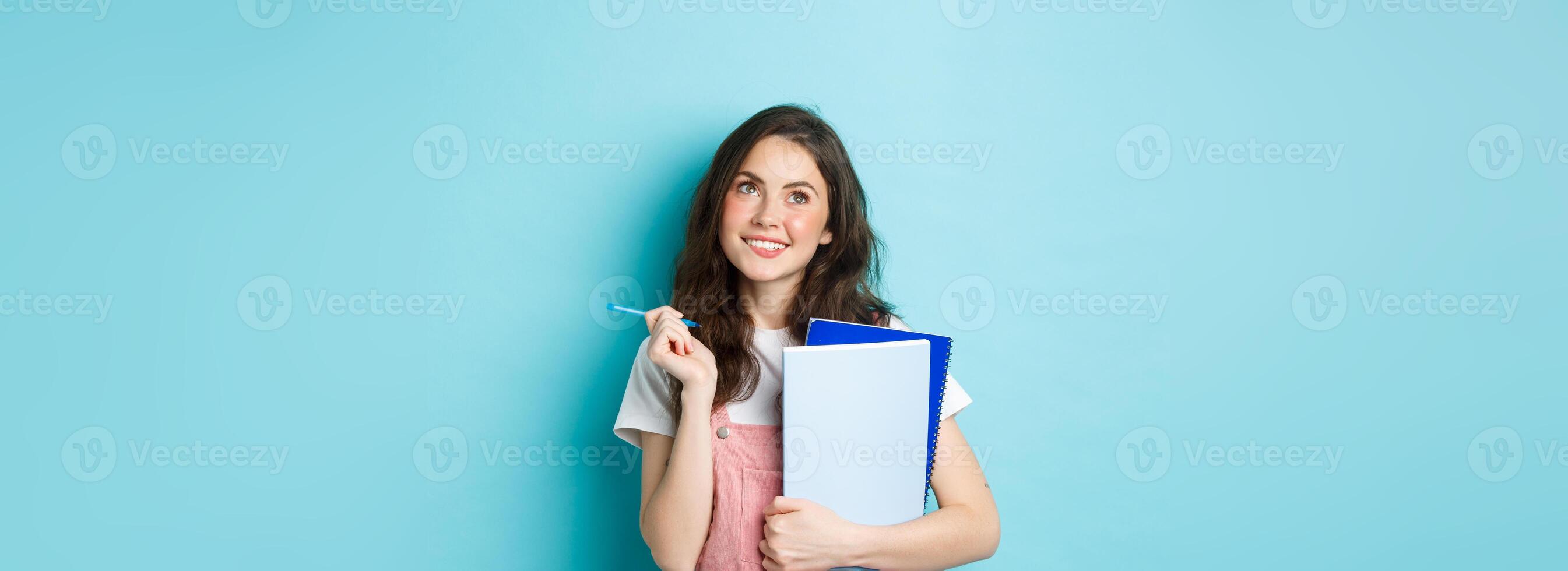 Beautiful girl student looking thoughtful at top copy space, standing with notebooks homework and smiling pensive, standing against blue background photo