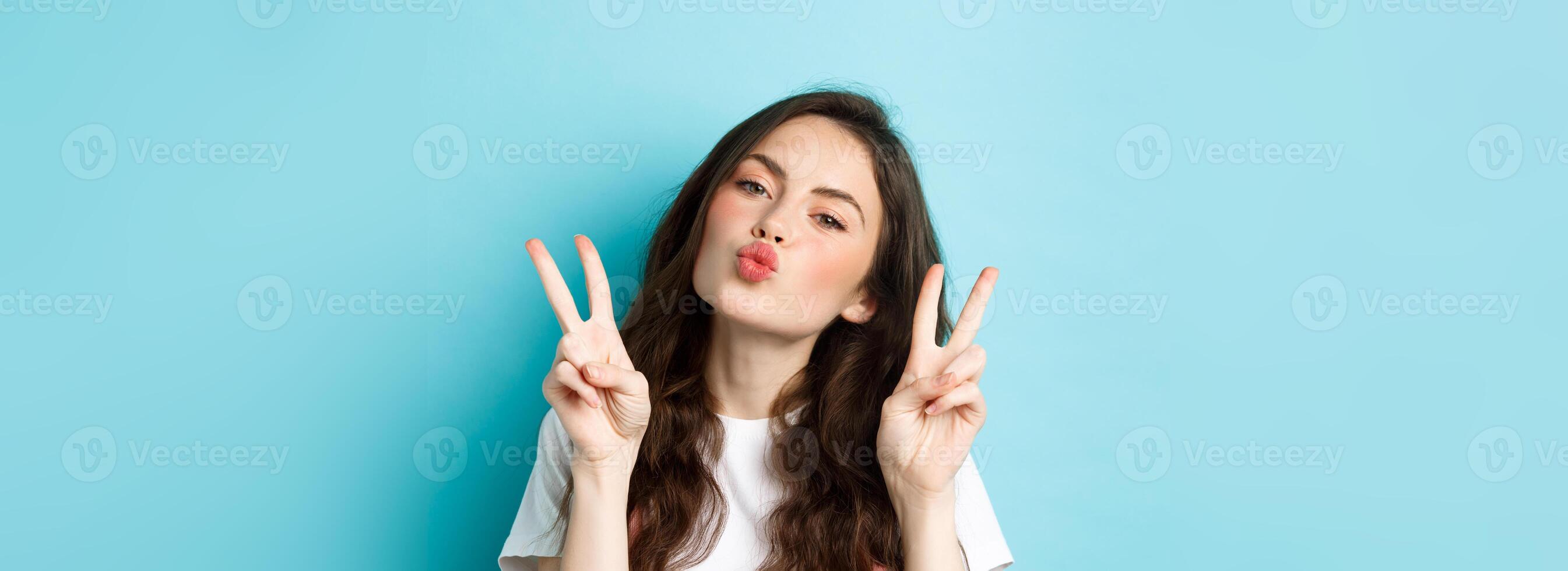 Close up portrait of glamour brunette girl making kissing face, showing kawaii v-sign and smiling cute at camera, standing happy against blue background photo